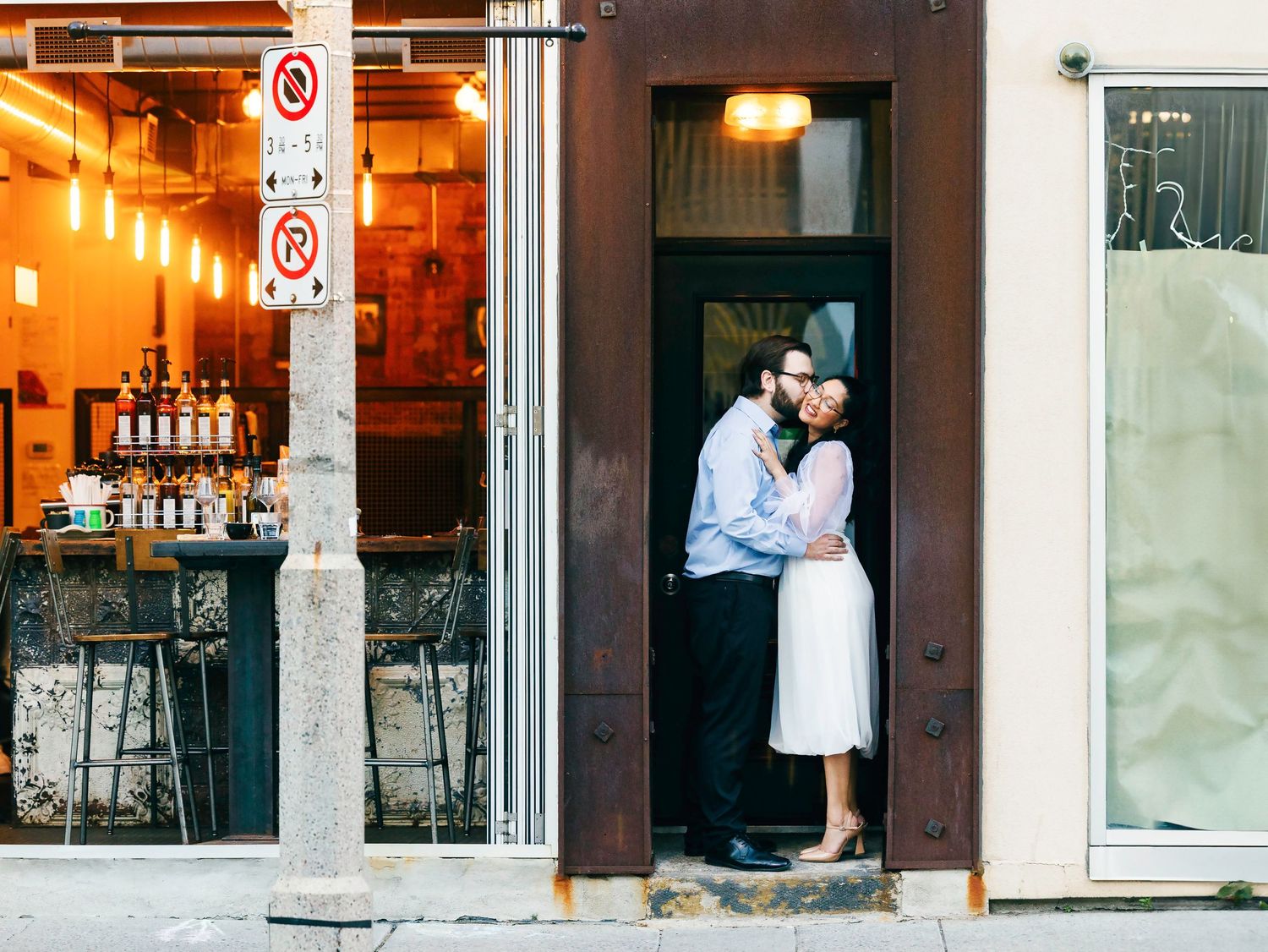 Couple sharing a romantic kiss in a charming city doorway beside a warmly lit restaurant storefront.