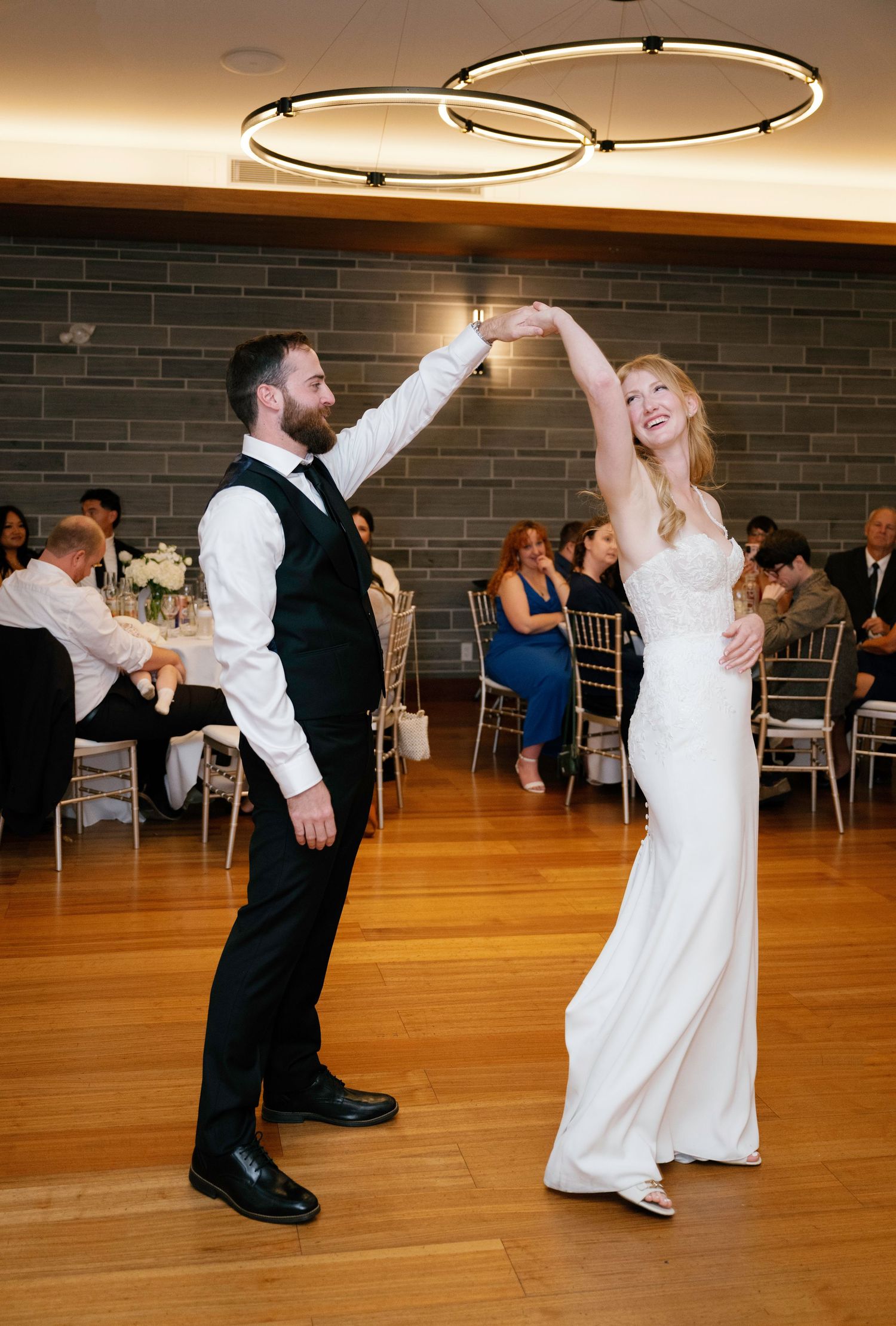 A couple dances together on a hardwood floor at a wedding reception beneath modern circular ceiling lights.