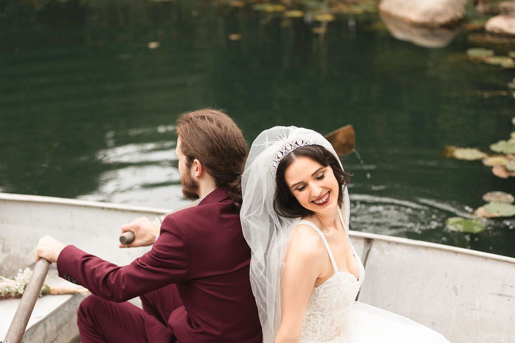 Bride and groom sitting back-to-back in a rowboat on a serene green lake, bride wearing veil and groom in burgundy suit.