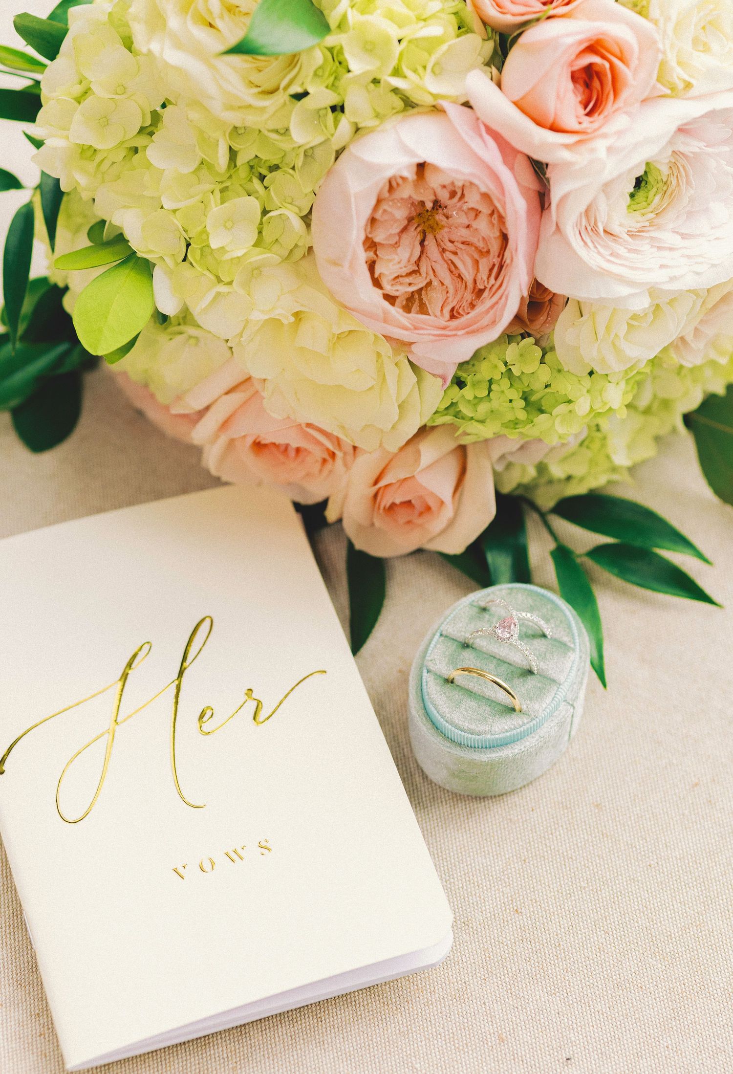Wedding flat lay with pink roses, hydrangeas, a vow book, and a diamond ring in a mint velvet box.