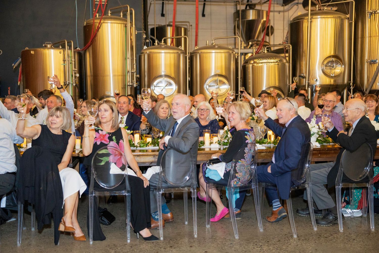 Guests dining and raising glasses at a brewery event surrounded by large golden fermentation tanks.