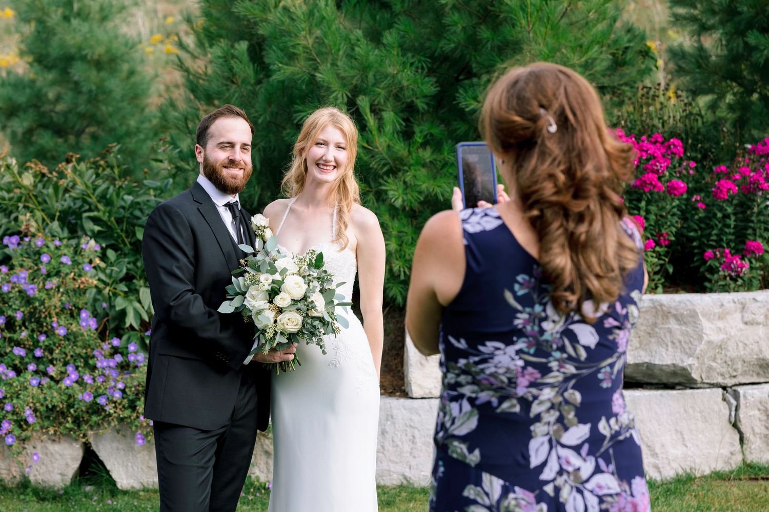 A wedding party in navy blue and white attire poses together in a garden with vibrant pink flowers.