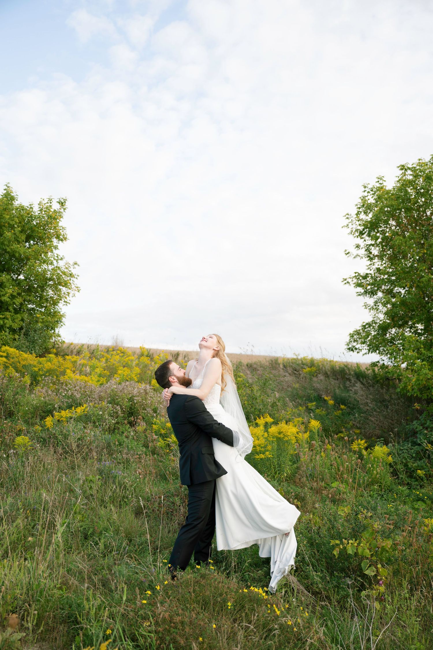 A couple in wedding attire dances joyfully in a wildflower meadow on a sunny afternoon.