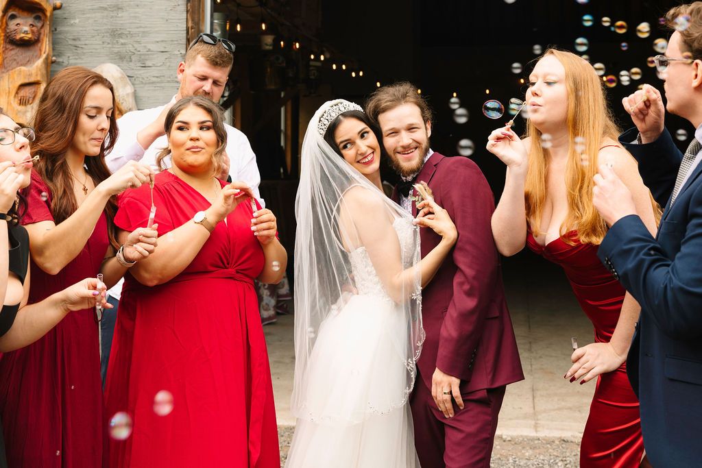 Bride in white gown and veil blowing bubbles with wedding party in red and burgundy attire at elegant venue.