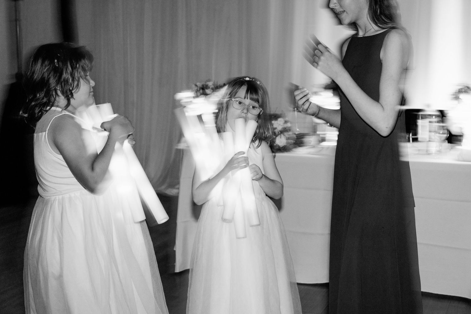 Black and white photo of flower girls in white dresses and a woman in a dark gown drinking from cups at an event.