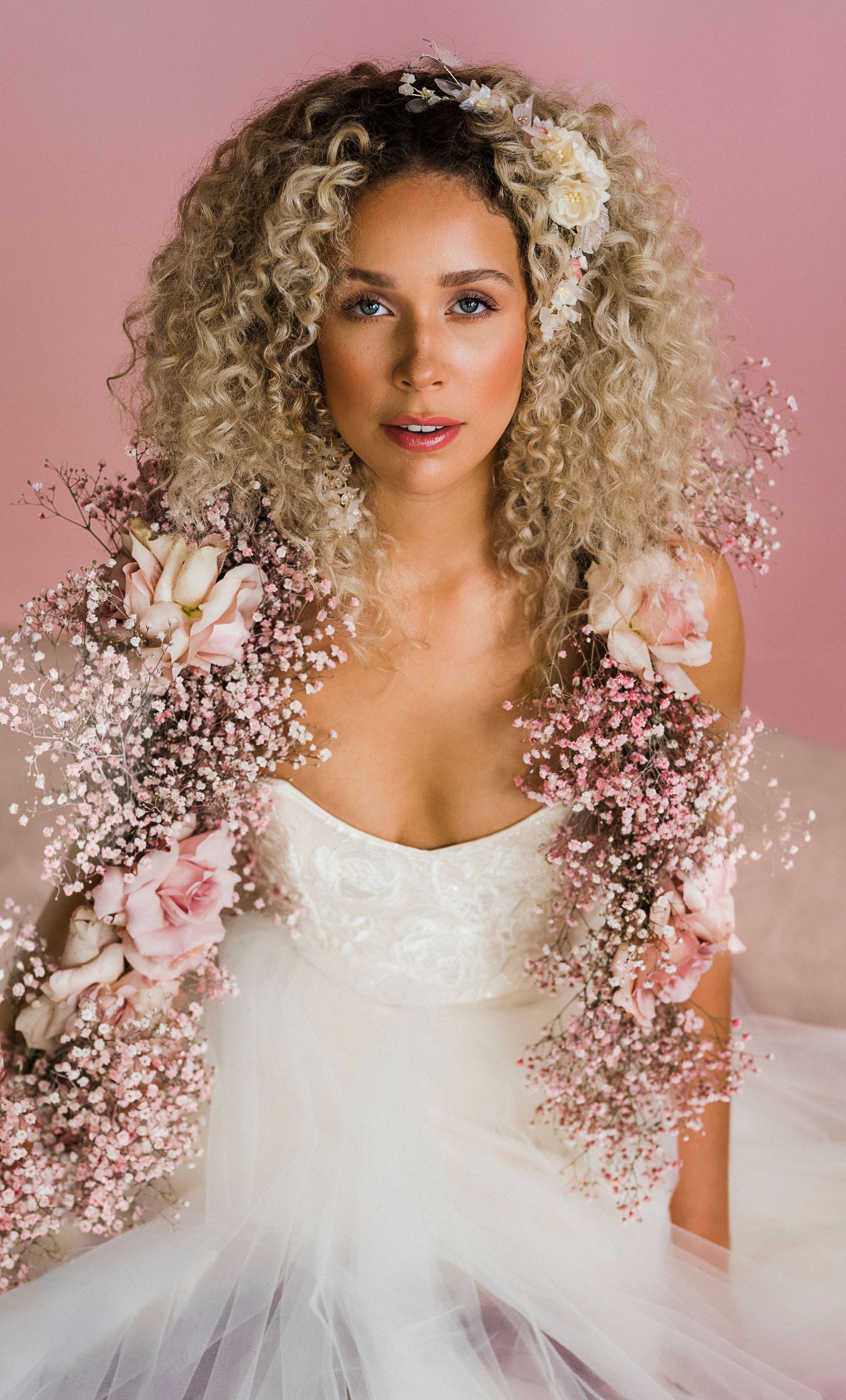 Bride in white dress with curly blonde hair adorned with baby's breath flowers against pink backdrop.