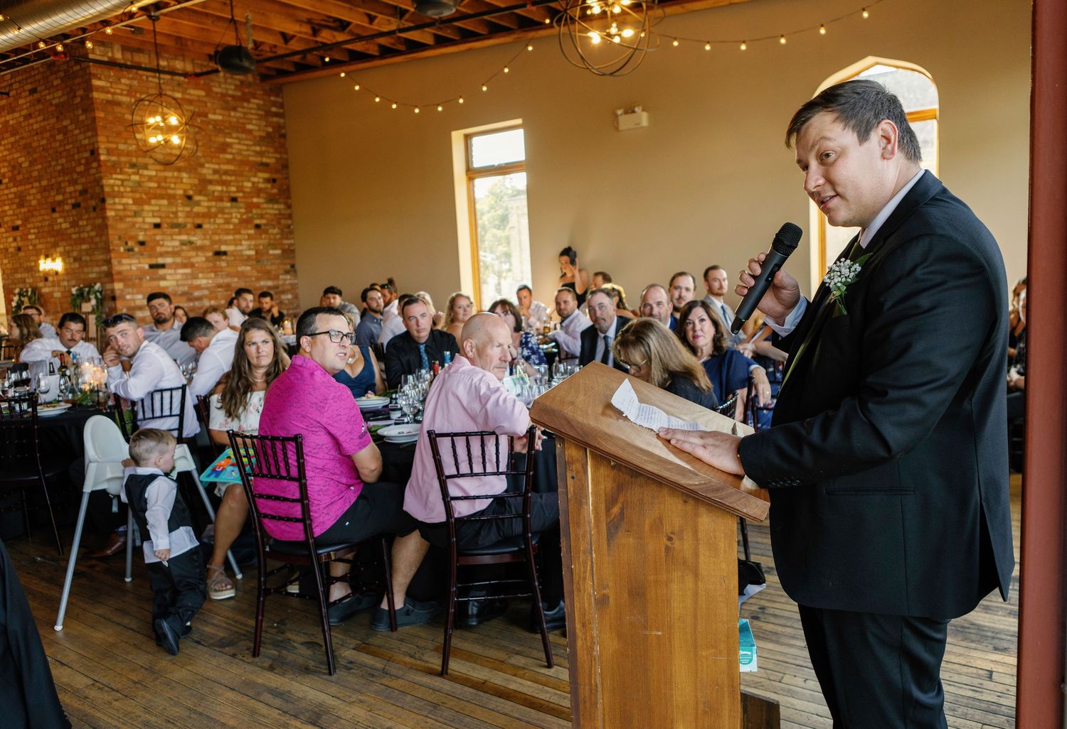 Wedding guests enjoy a reception in a rustic brick-walled venue with string lights and elegant table settings.