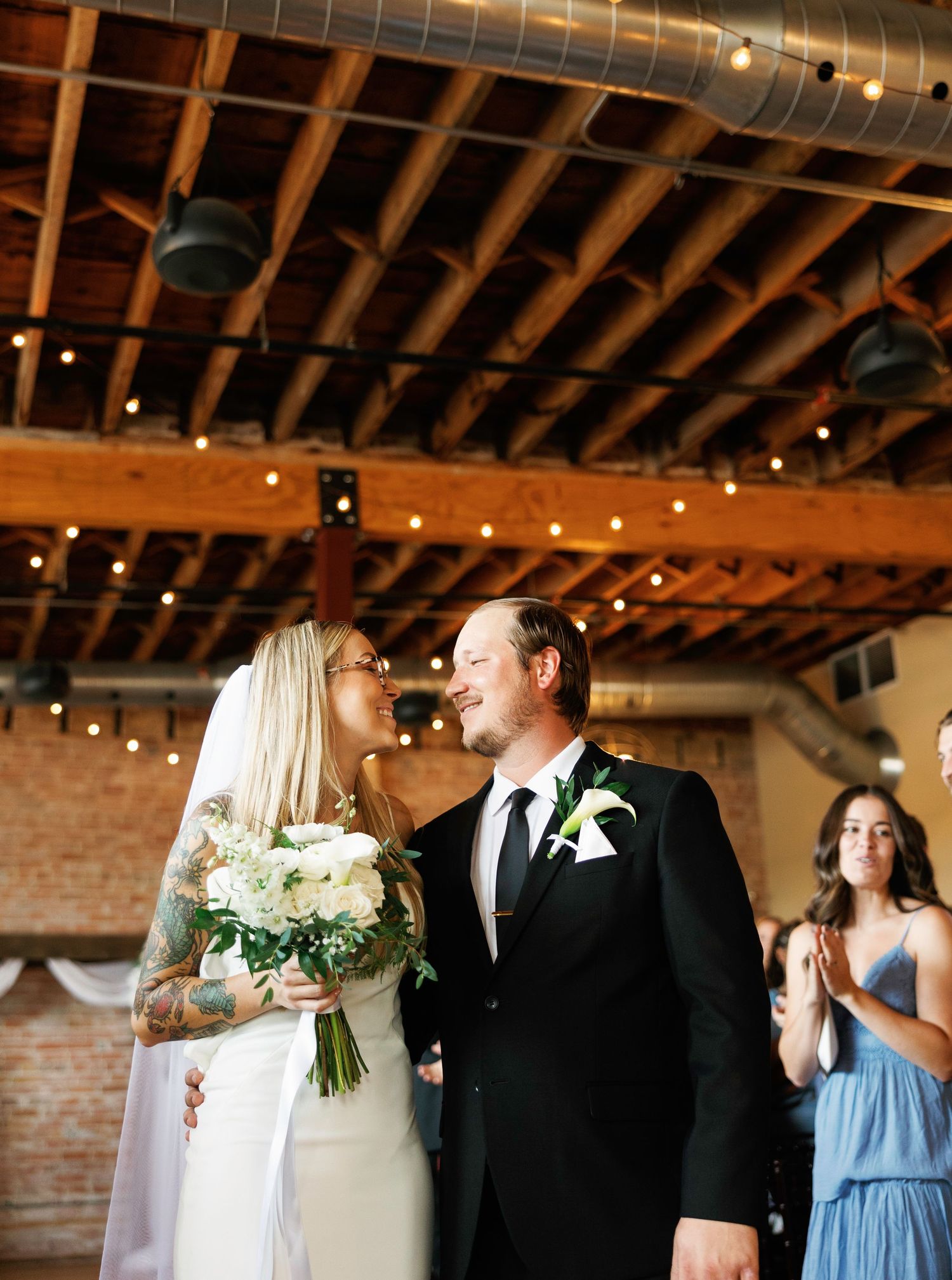 A couple shares an intimate moment during their wedding ceremony in a rustic brick-walled venue with string lights above.