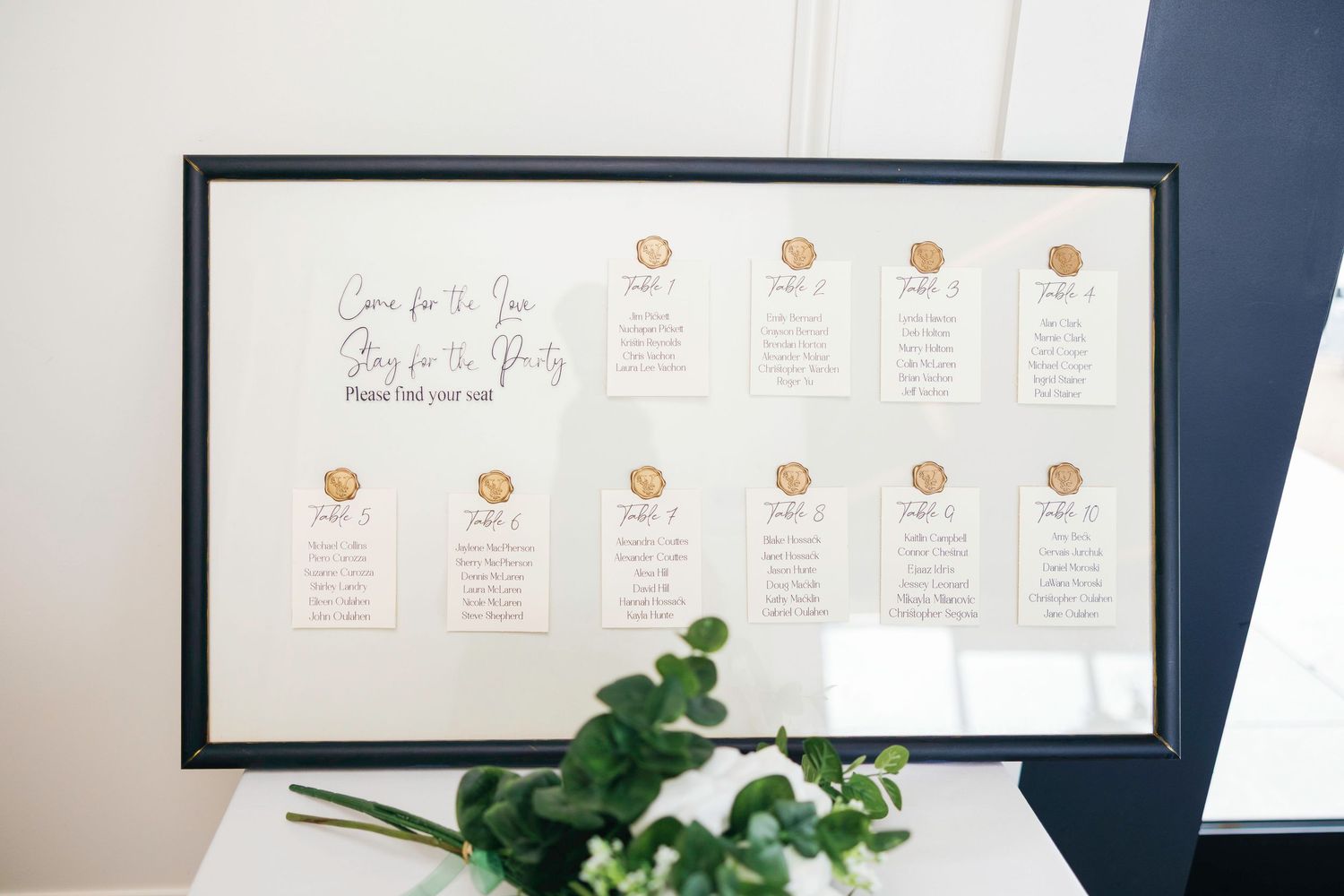 Modern minimalist wedding seating chart displayed on navy blue wall with greenery accents.