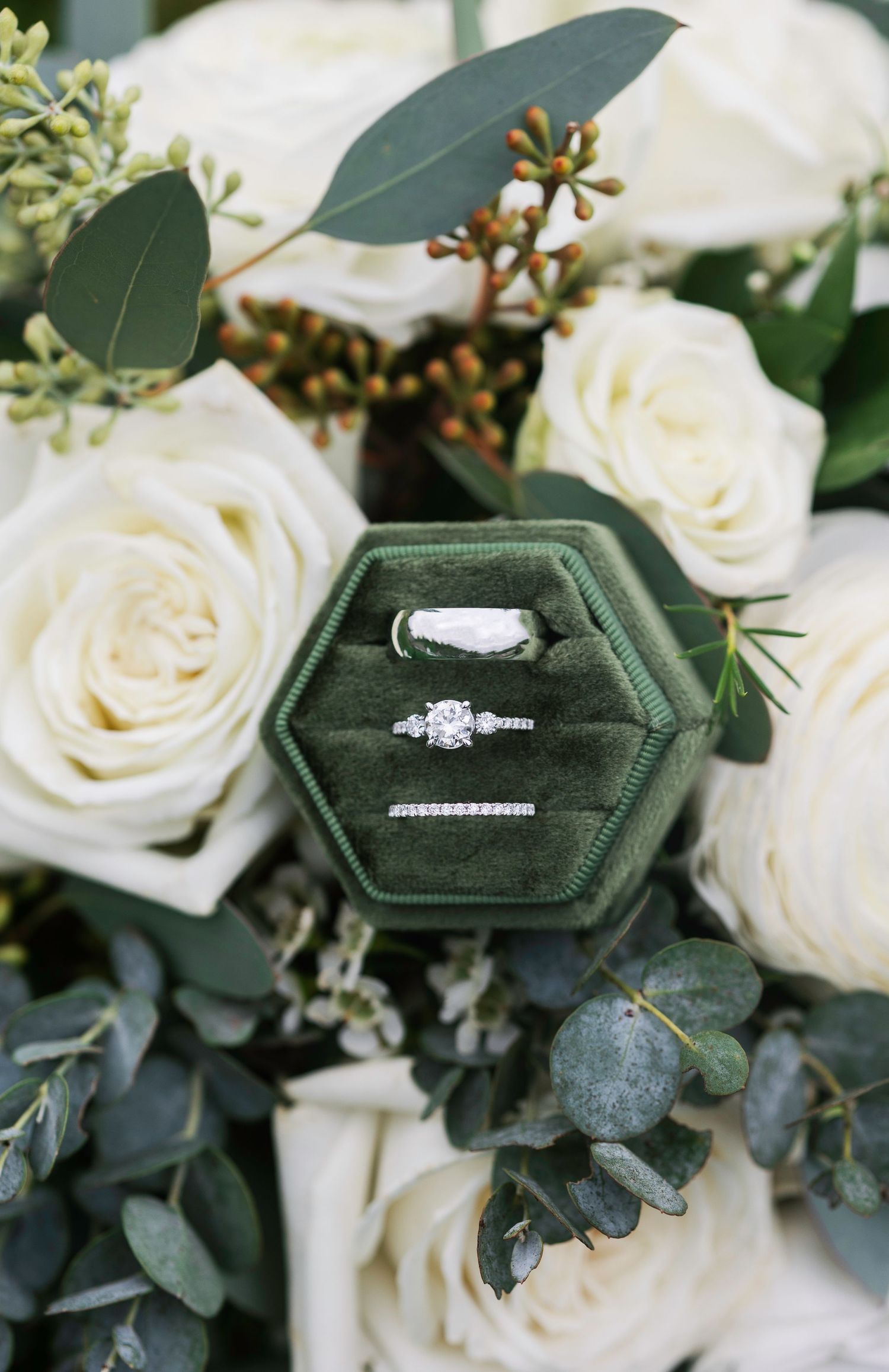 Wedding rings displayed on dark green velvet hexagonal ring boxes with white roses.