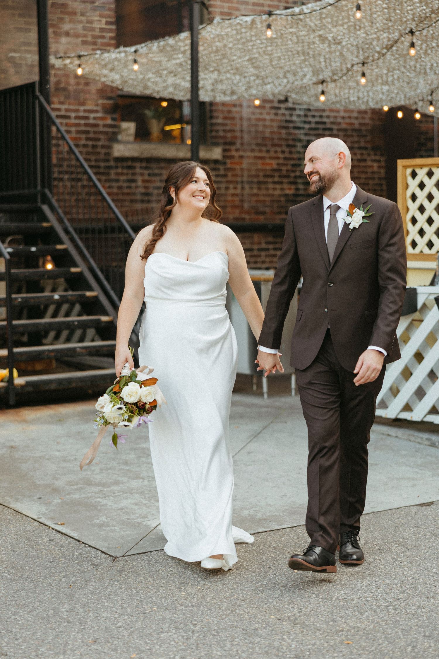 A joyful wedding couple walking together on an urban patio with string lights and brick walls.