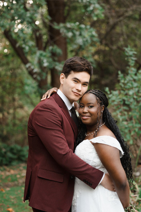 A romantic outdoor wedding portrait of a couple in burgundy suit and white dress among trees.