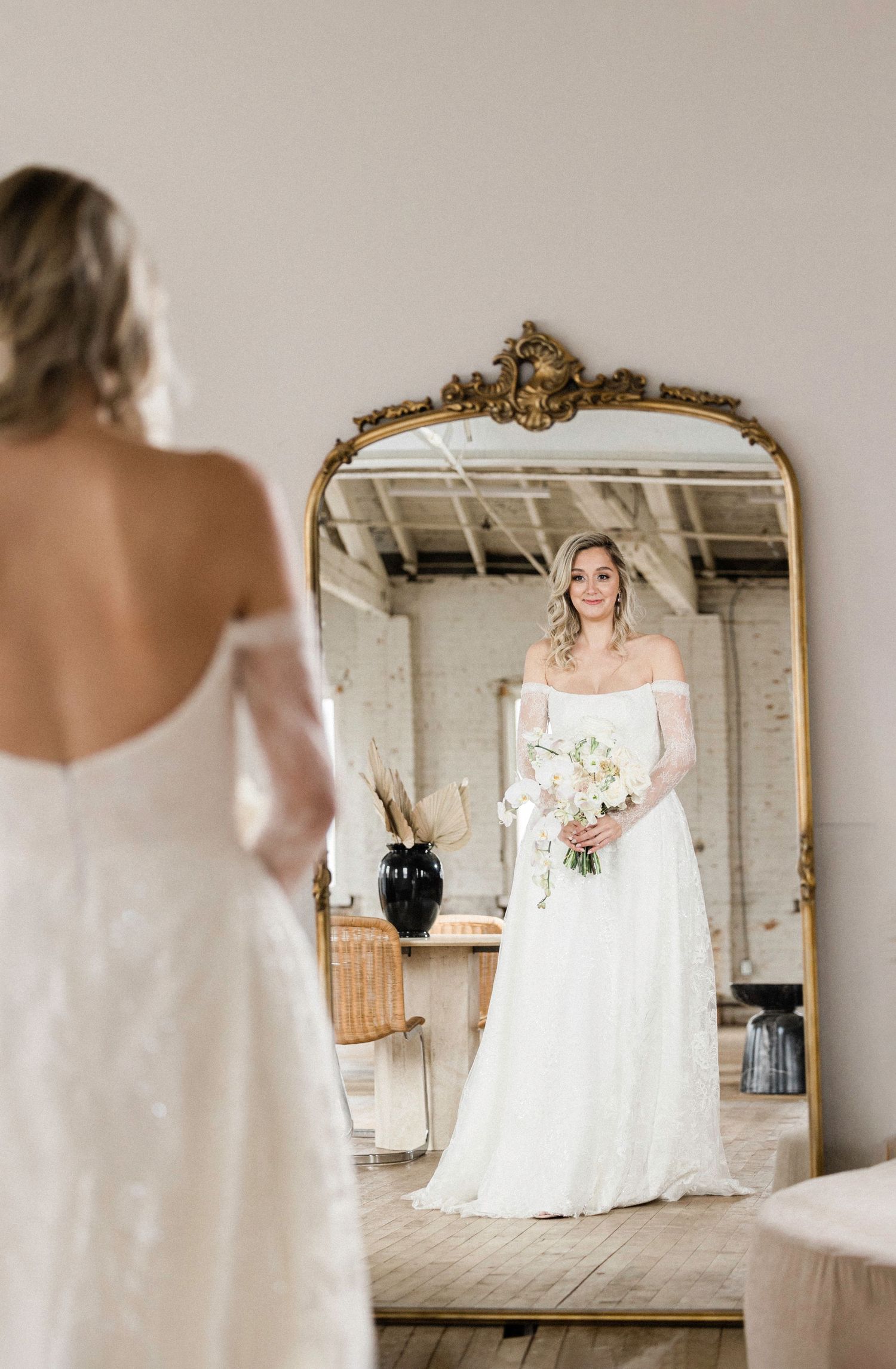 Bride in white off-shoulder gown holding bouquet, reflected in ornate gold mirror in rustic studio.