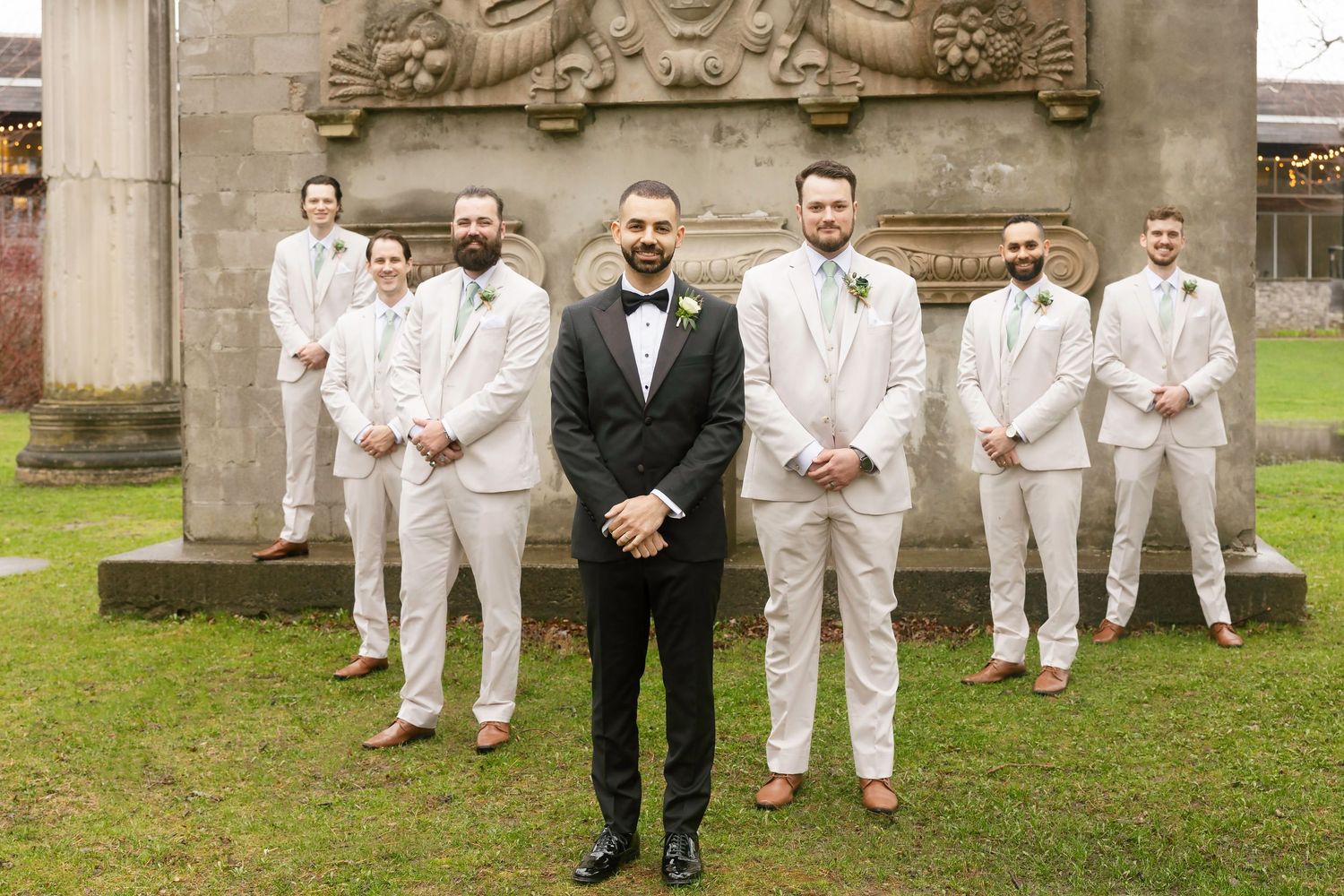 Groom in black tuxedo stands before groomsmen in white suits near stone column at outdoor wedding venue.