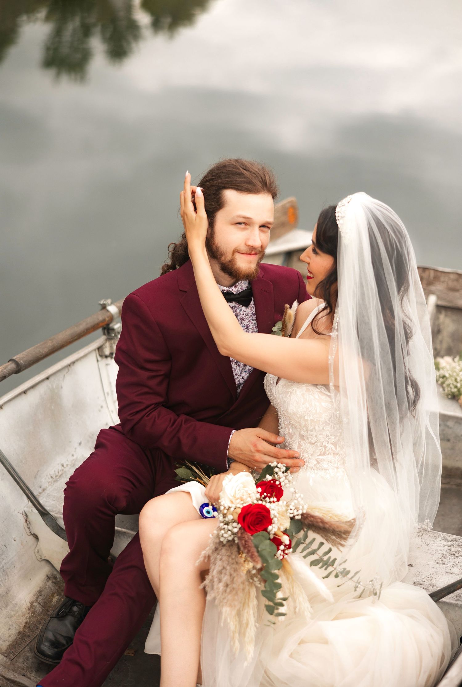 Newlywed couple sitting in a boat, groom in burgundy suit smiling, bride in white gown with veil holding red bouquet.