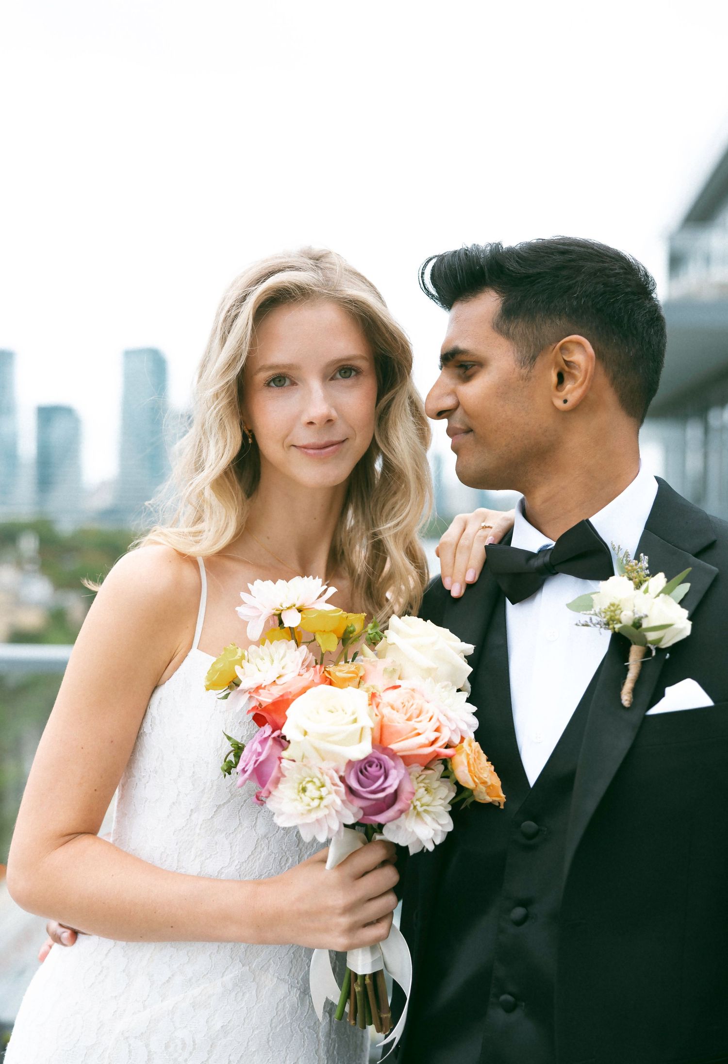 Bride holding colorful bouquet with groom in black tuxedo posing on rooftop with city skyline backdrop.