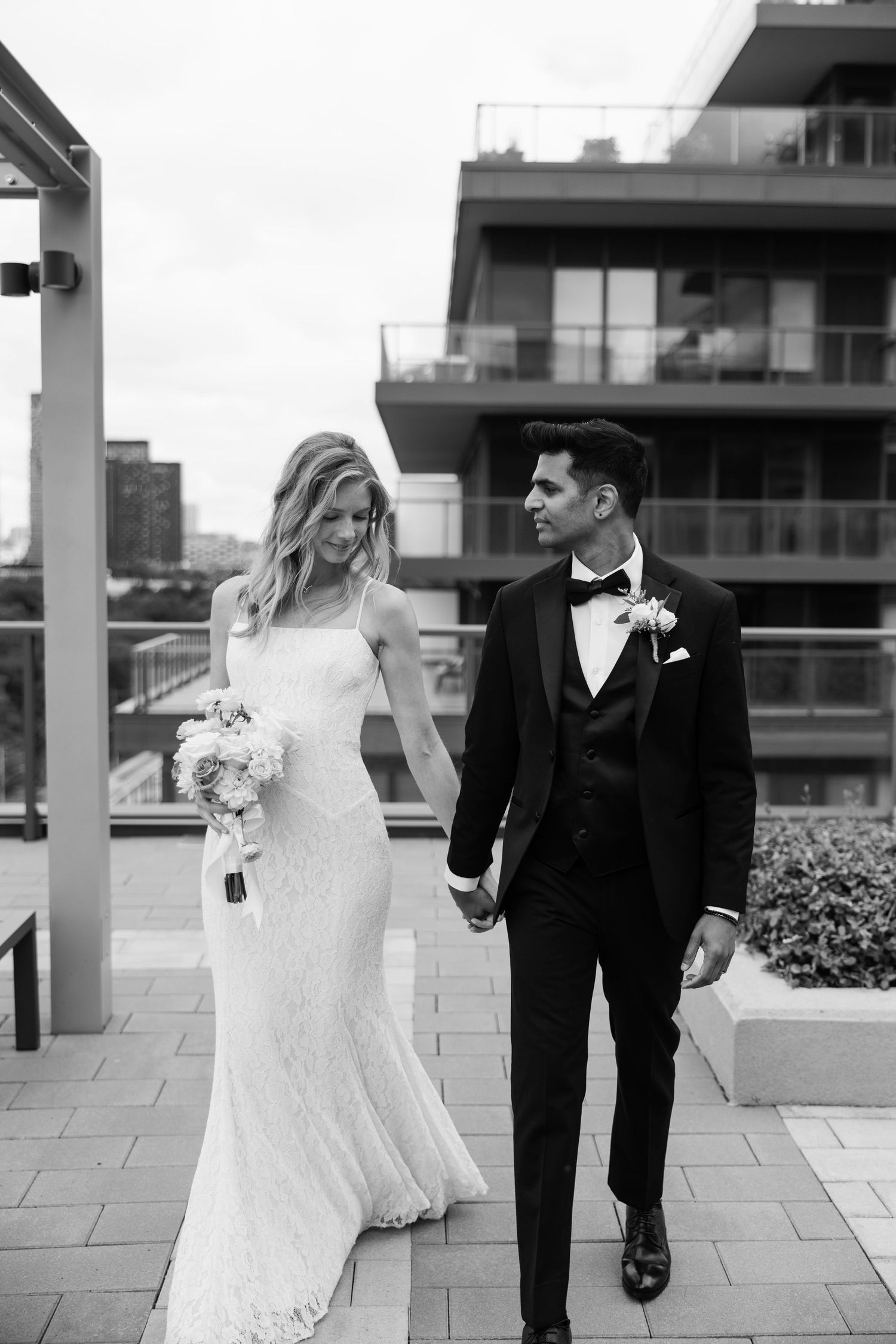 Black and white photo of a bride and groom holding hands on a rooftop terrace, with modern building in background.