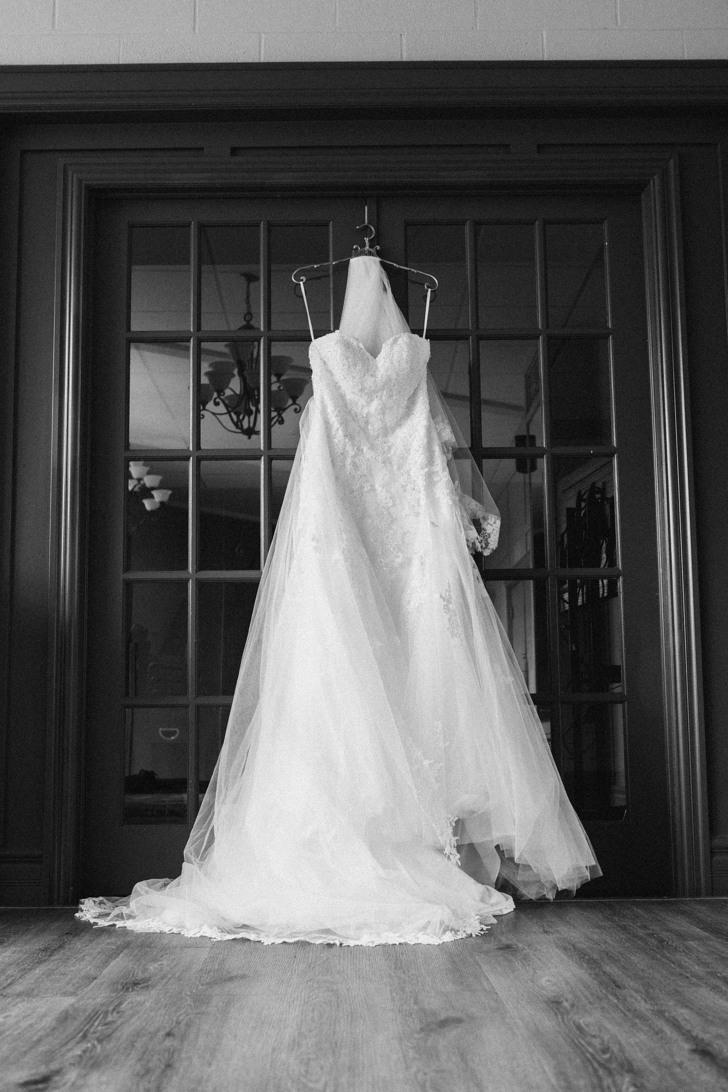 Black and white photo of an elegant lace wedding dress with veil hanging on a hanger before french doors.