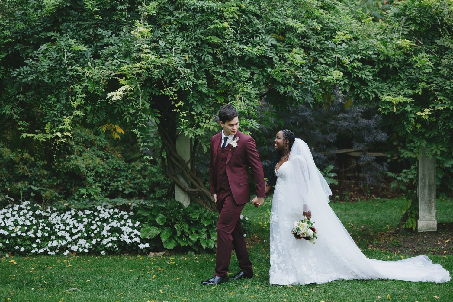Interracial couple in wedding attire holding hands in lush garden, groom in burgundy suit, bride in white gown with veil.