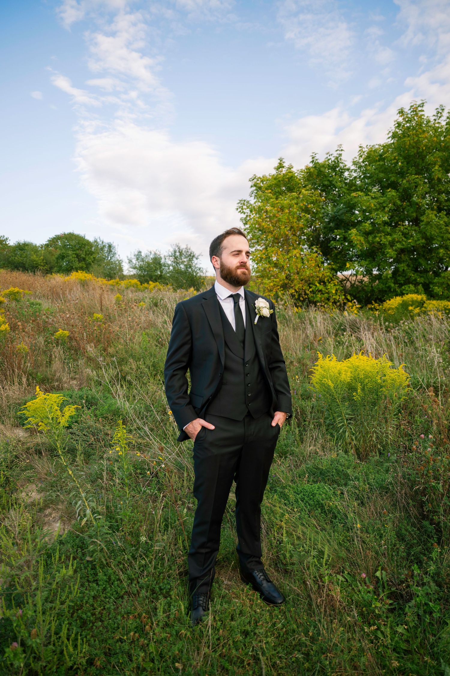 A person in a black formal suit poses in a field of tall grass and wildflowers on a sunny day.