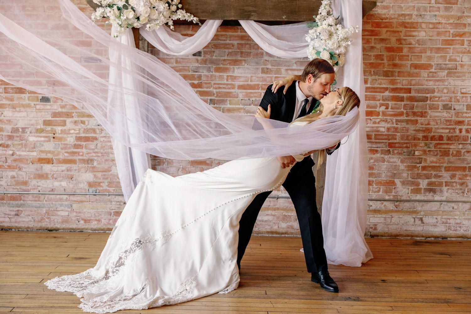 A couple in formal wedding attire dances together in a rustic brick-walled venue with flowing white drapery.