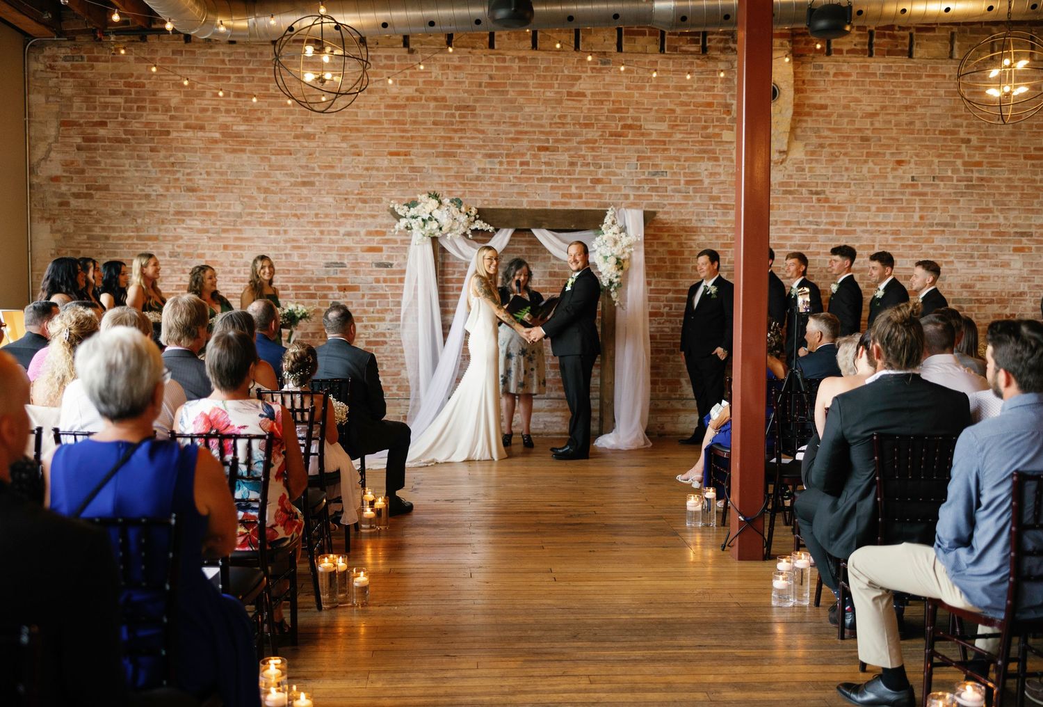 Rustic wedding ceremony setup with exposed brick walls, wooden beams, and hanging string lights.