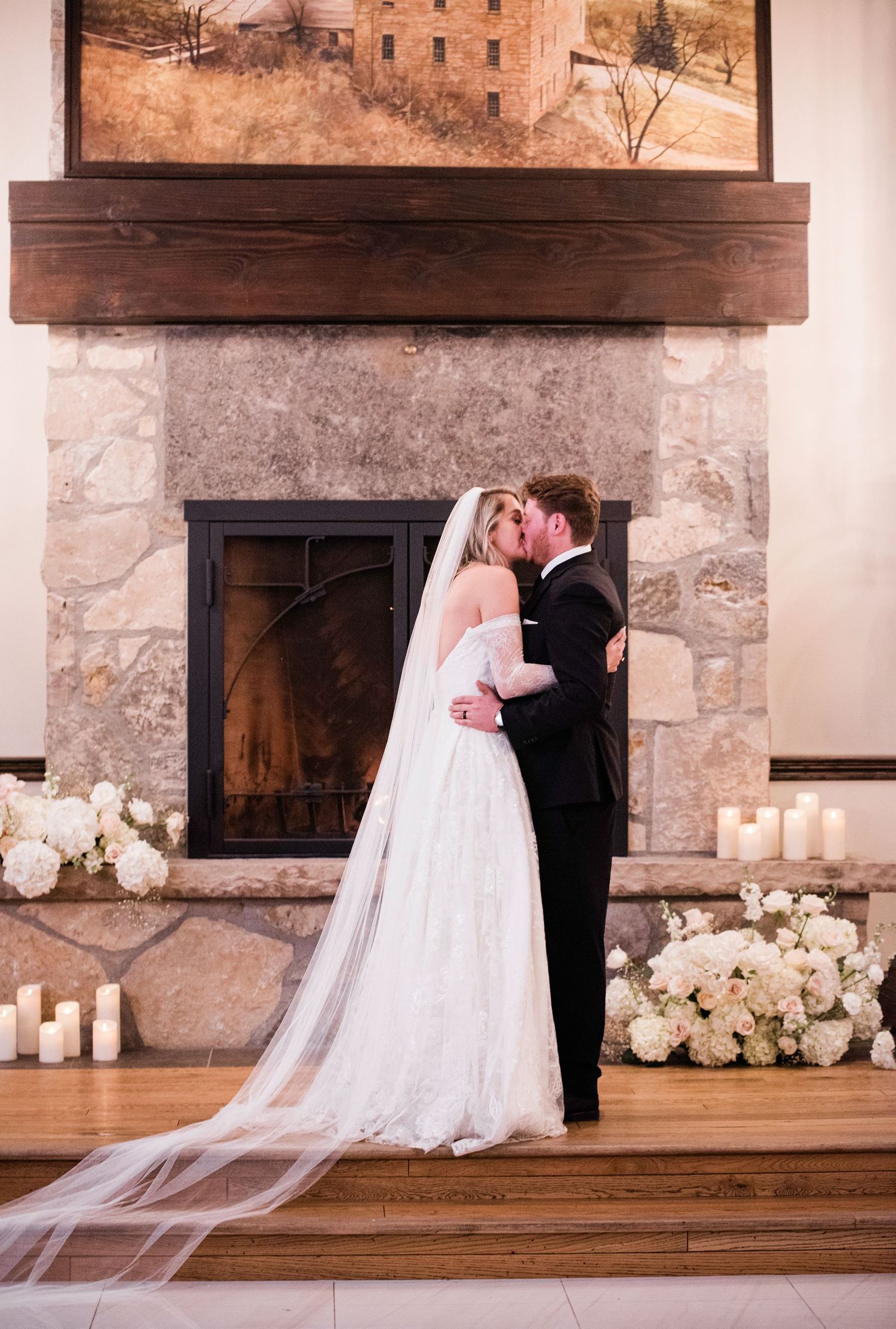 Wedding couple poses in front of rustic stone fireplace decorated with white floral arrangements.