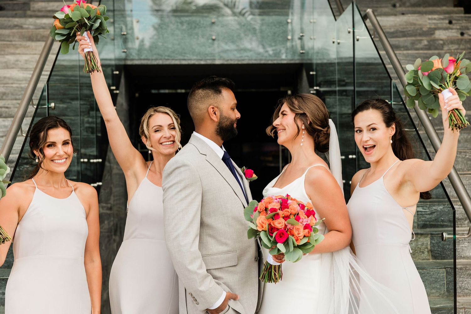 Wedding party celebrating on modern glass staircase with coral and pink floral bouquets.