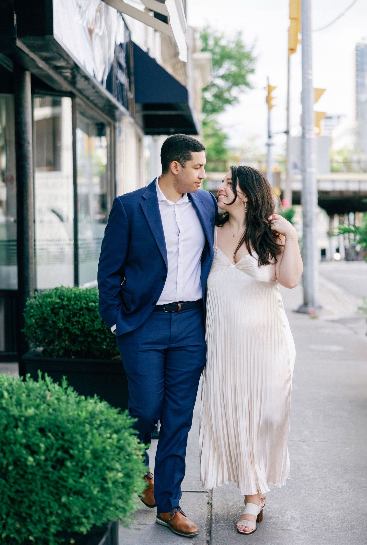 Couple in elegant attire poses on urban sidewalk, man in navy suit and woman in white pleated dress.