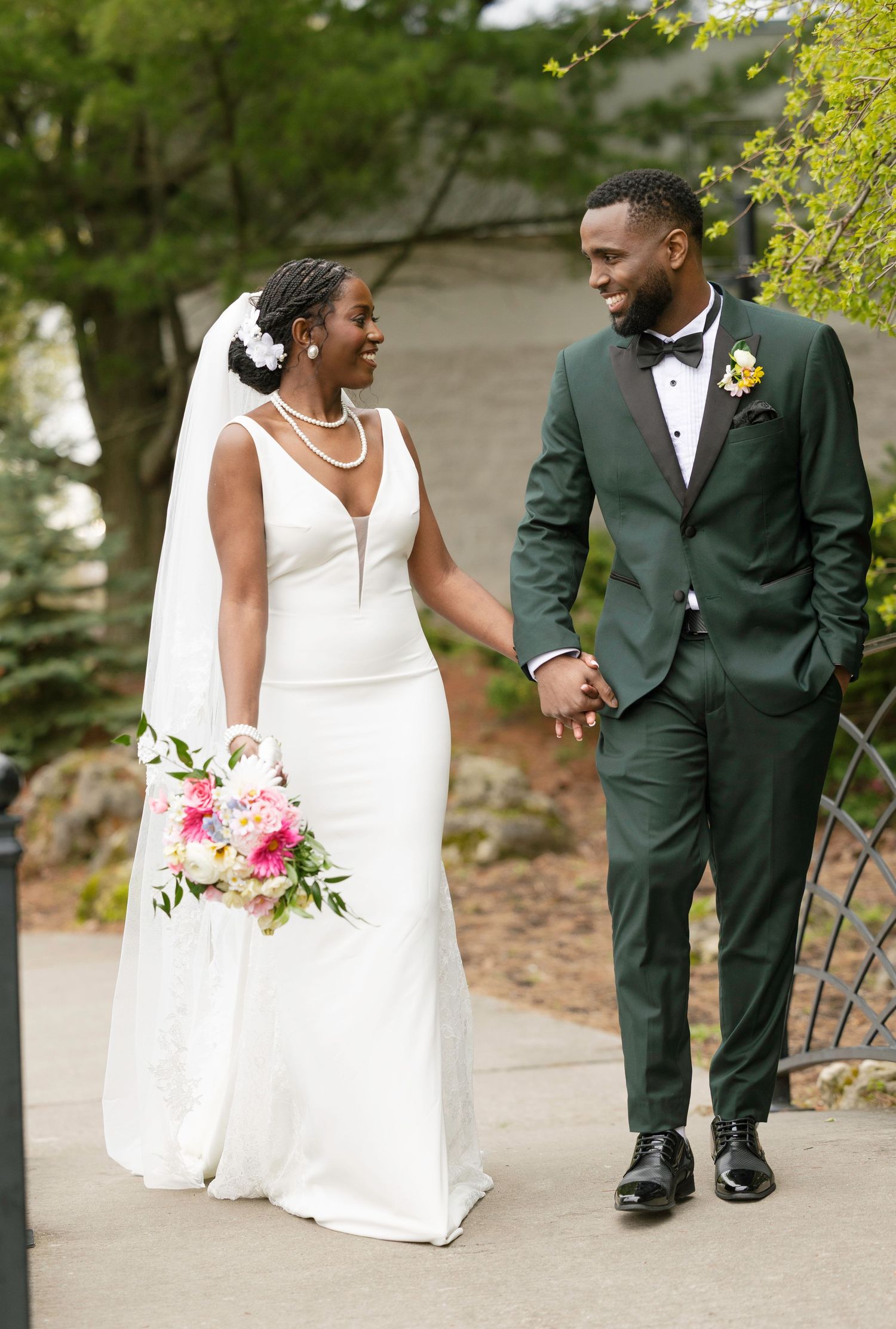 A bride in a white gown and groom in a dark green tuxedo hold hands and smile at each other outdoors.