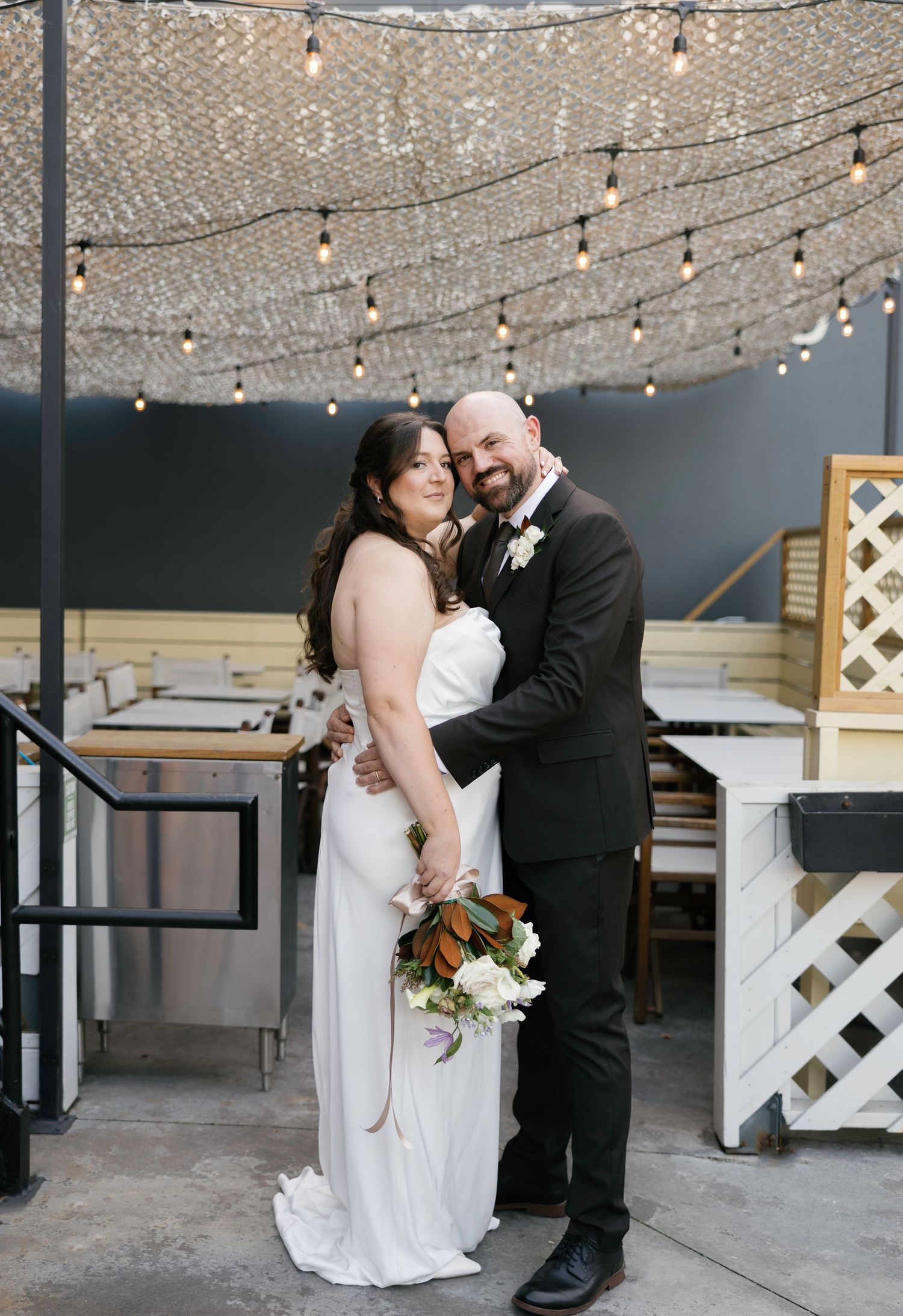 Bride and groom embrace under string lights at an indoor wedding venue, bride holding a white floral bouquet.