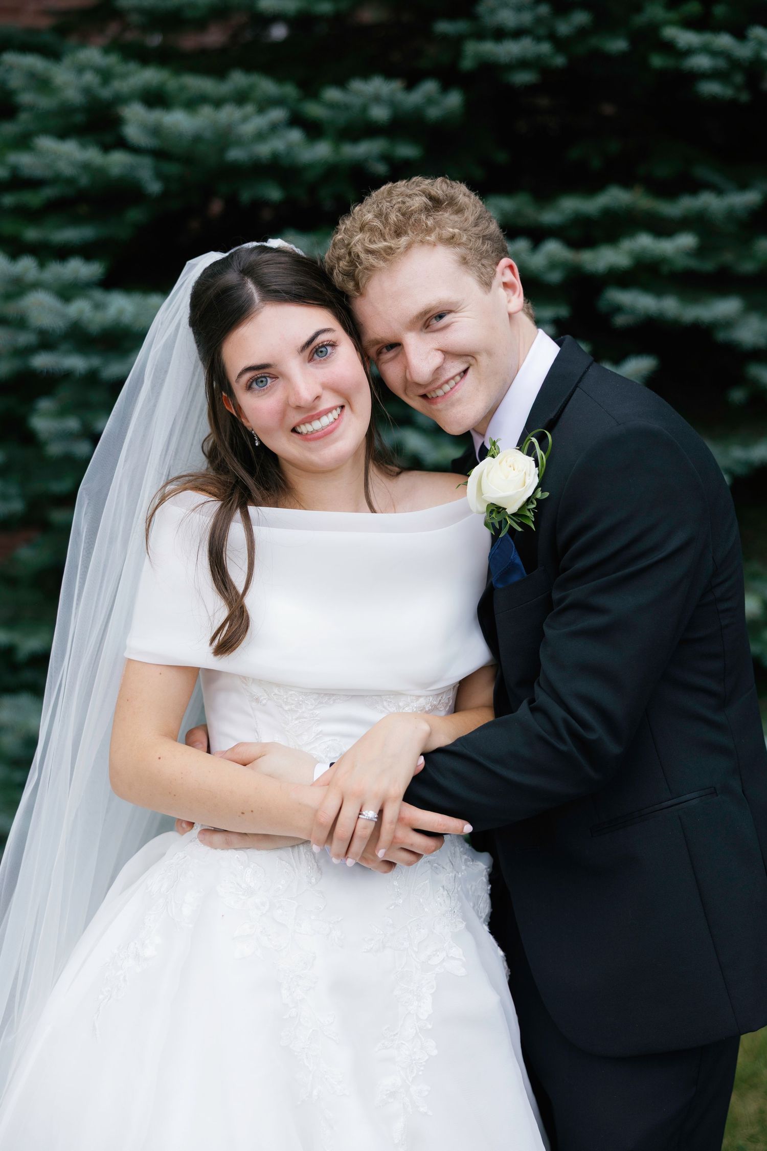 Smiling bride and groom embrace outdoors, bride in white gown with veil, groom in black suit with boutonniere.