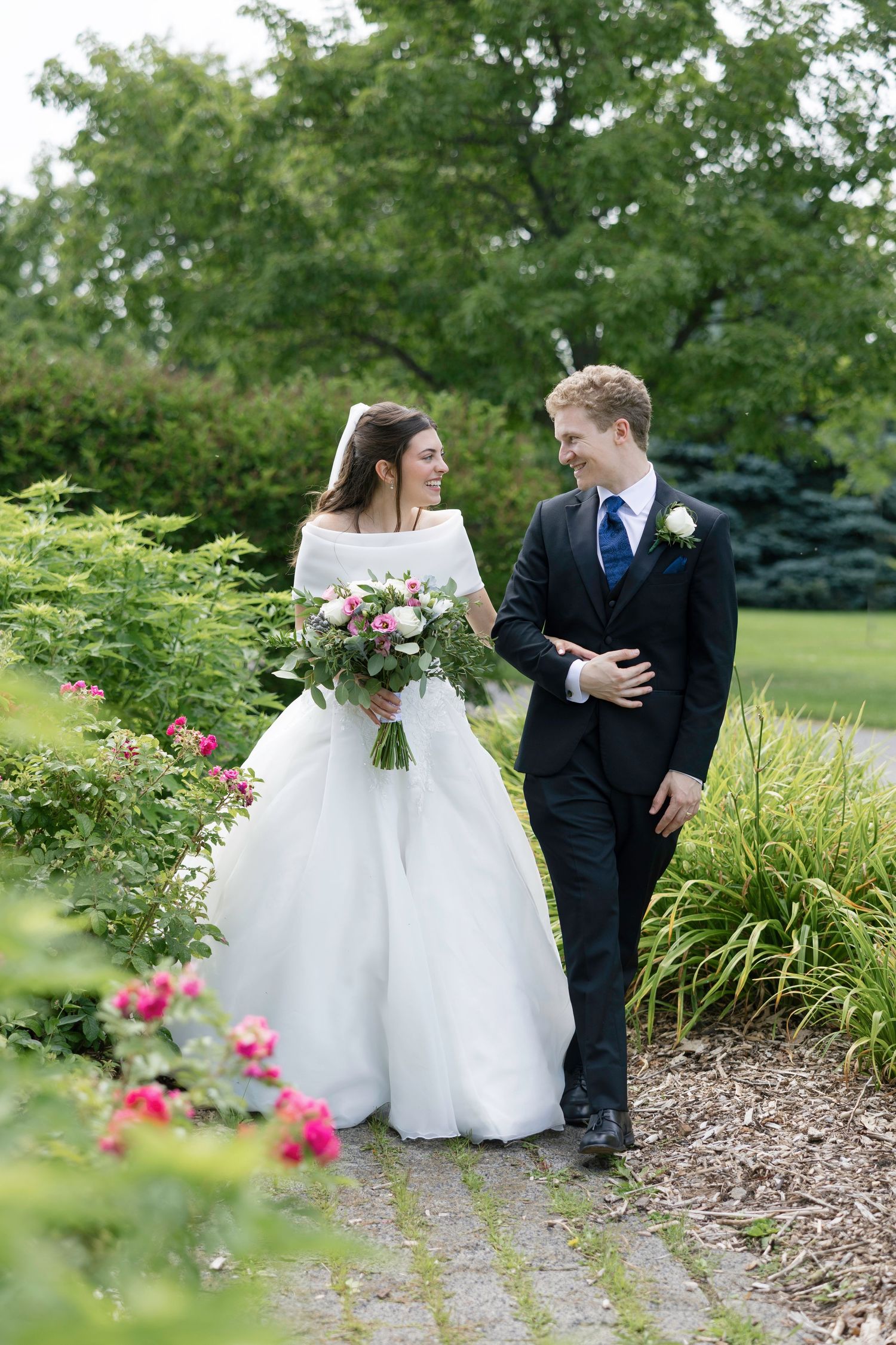 Bride and groom walking together on garden path, bride holding pink bouquet in white off-shoulder gown.