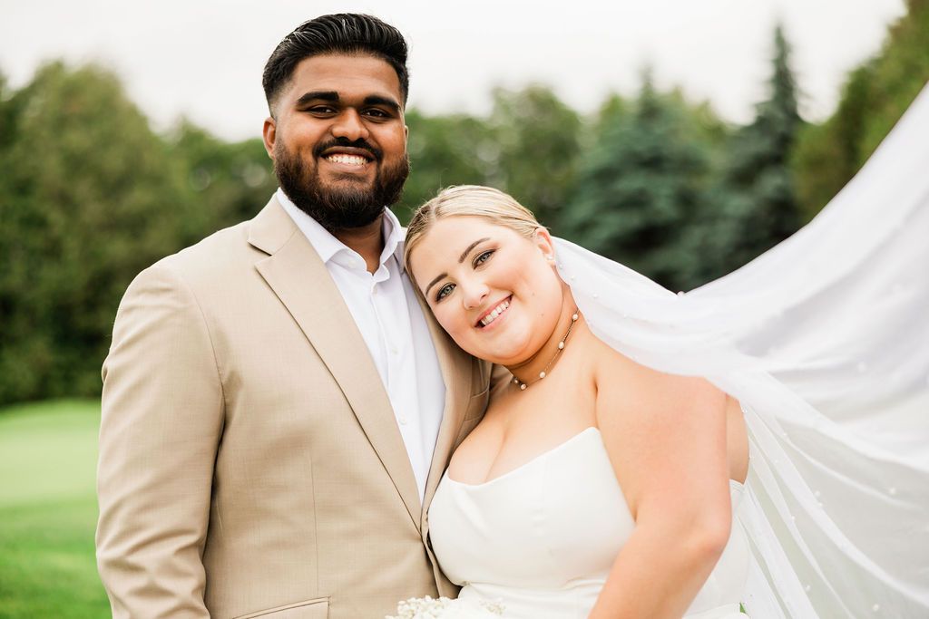 Smiling interracial couple in wedding attire outdoors, groom in tan suit and bride in white dress with flowing veil.