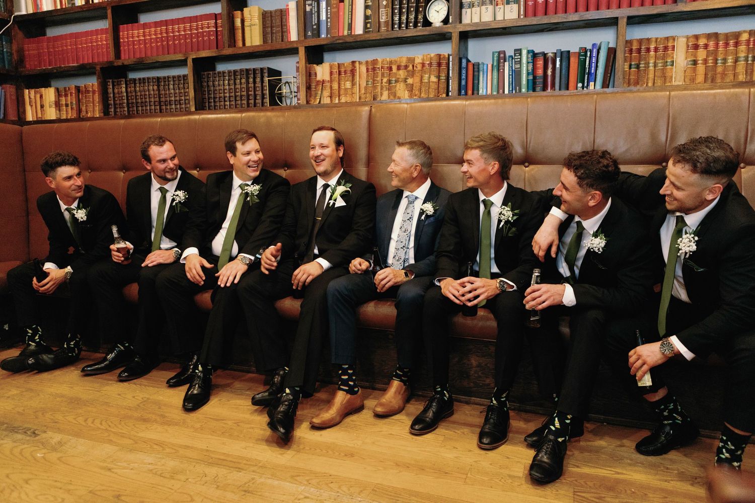 Groomsmen in dark suits with green ties laughing together on a leather sofa in a library setting.