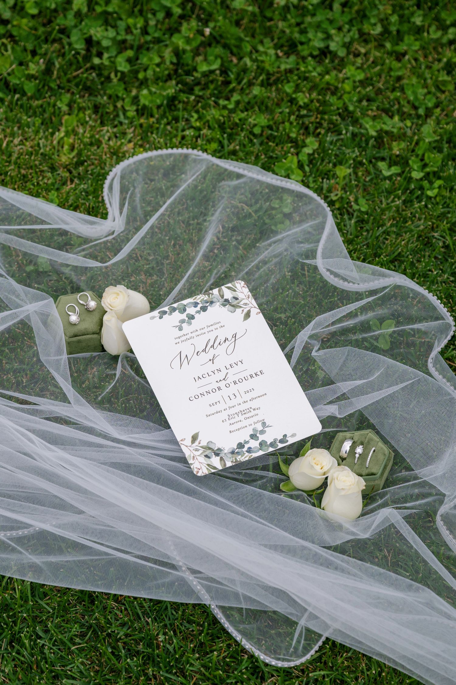 White wedding veil draped on green grass with delicate invitation cards and small white roses creating an elegant bridal detail.