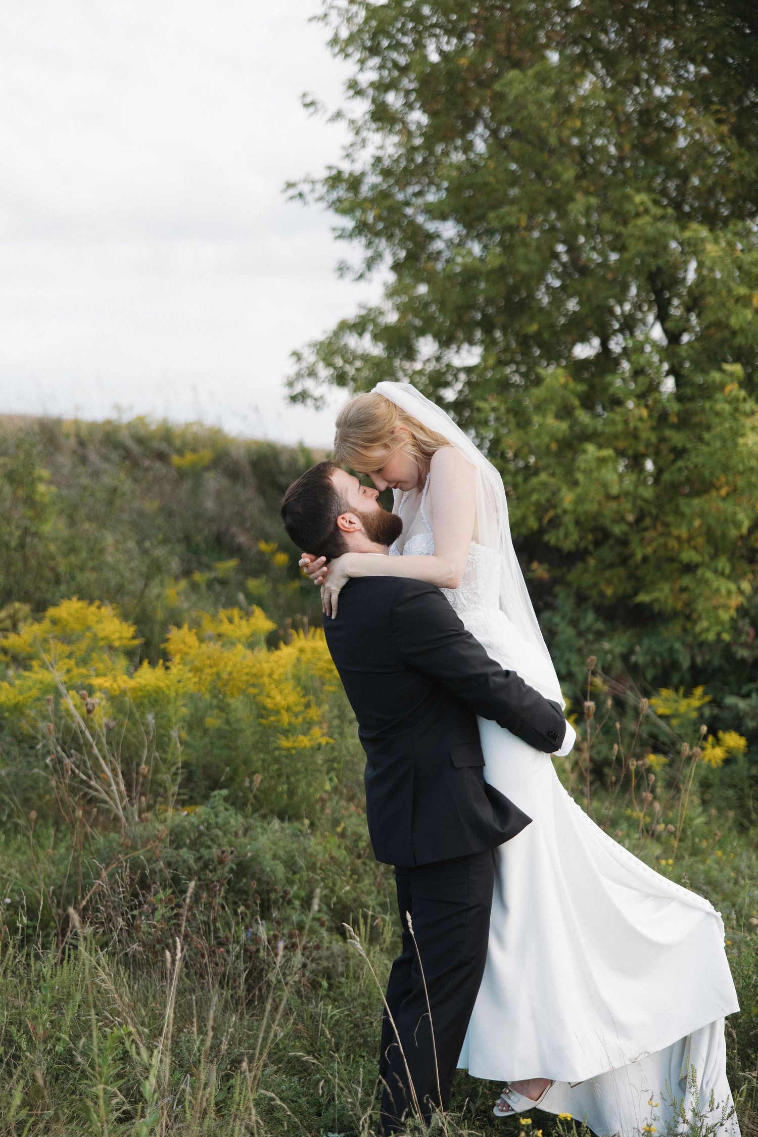 A couple in wedding attire embraces and dances in a field of wildflowers and autumn trees.