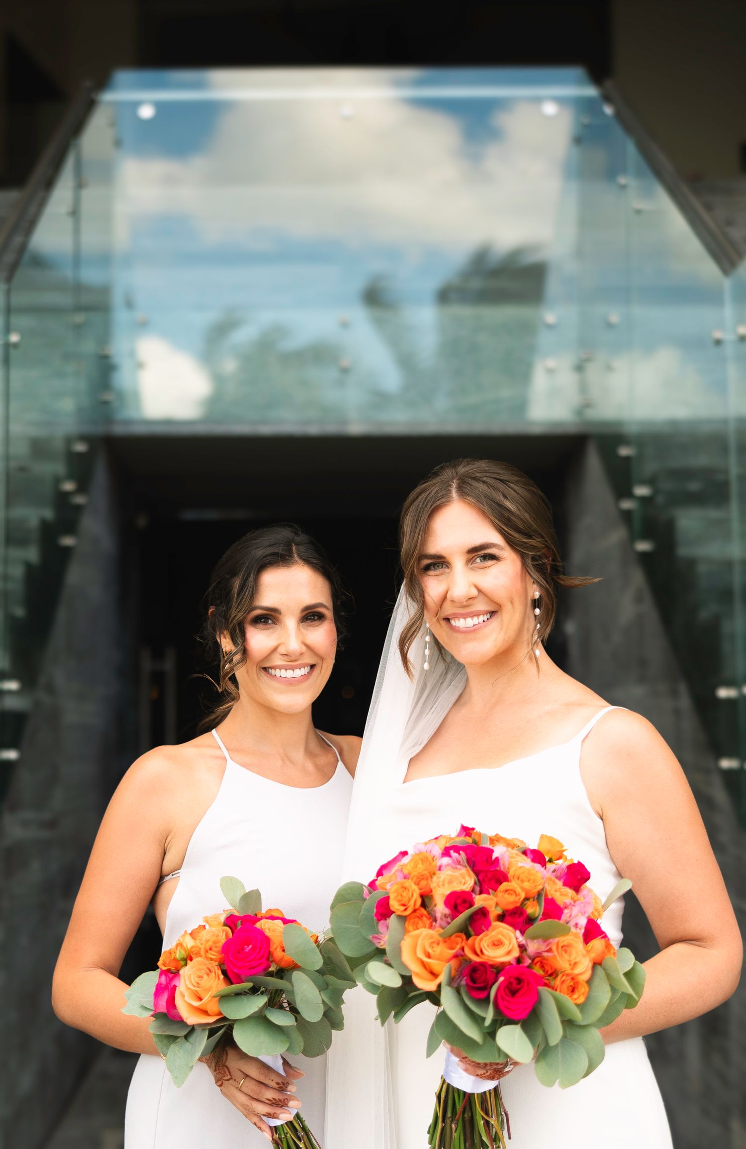 Bridesmaids in white dresses holding vibrant pink and orange bouquets pose together outdoors.