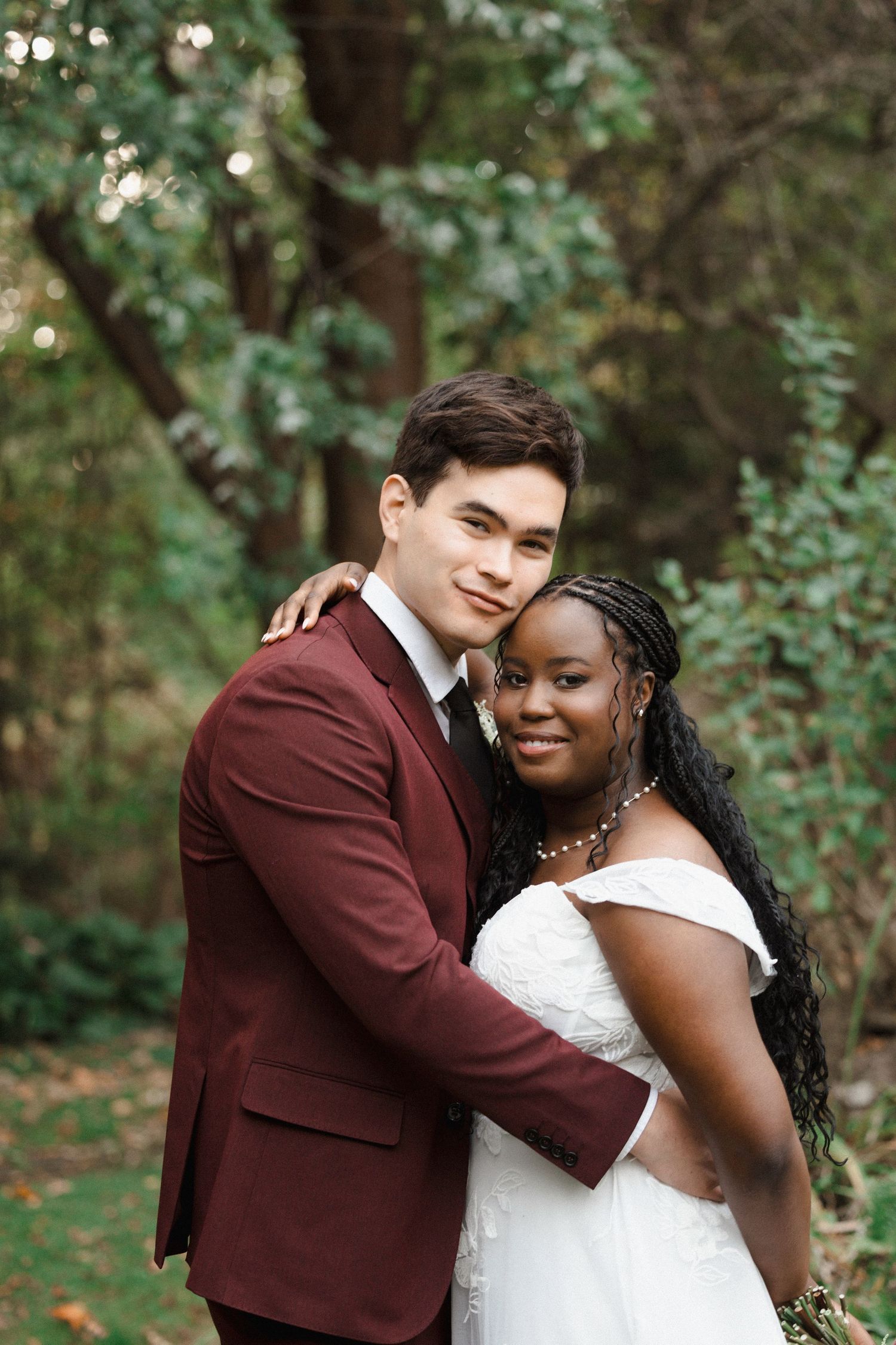 A happy couple embraces outdoors in wedding attire, with the groom in burgundy suit and bride in white dress.