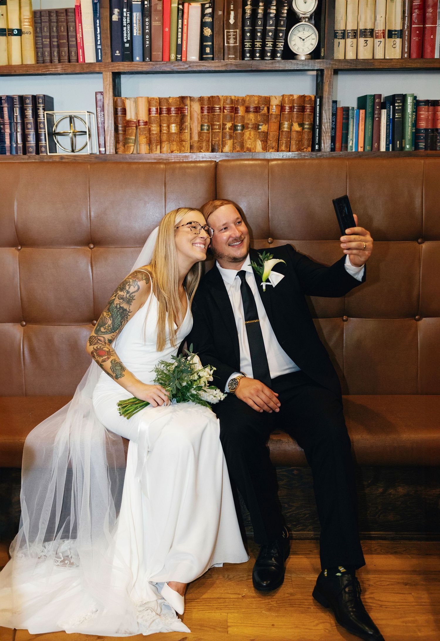 A bride and groom share intimate moments on a brown leather bench in a library setting with vintage books in the background.