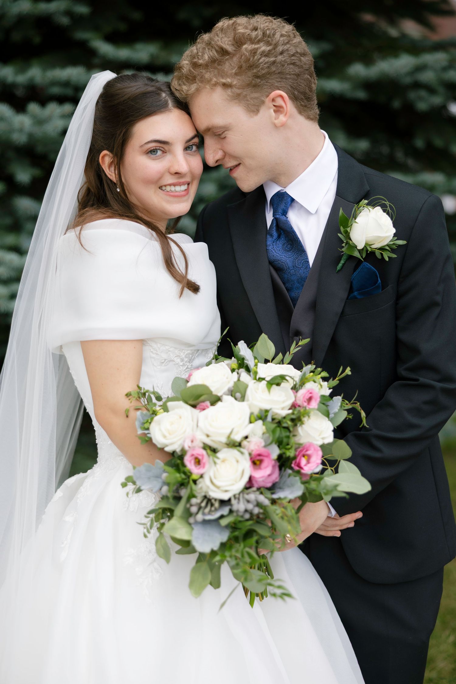 Bride and groom share a tender moment, holding a bouquet of white and pink roses on their wedding day.