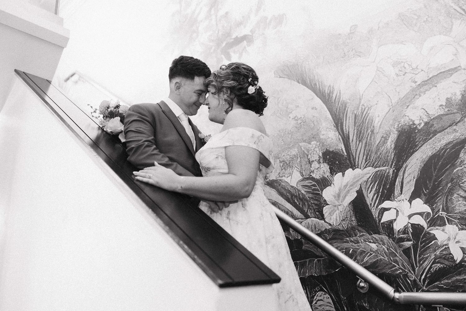 Black and white wedding photo of a couple sharing a romantic moment on a staircase with floral mural backdrop.