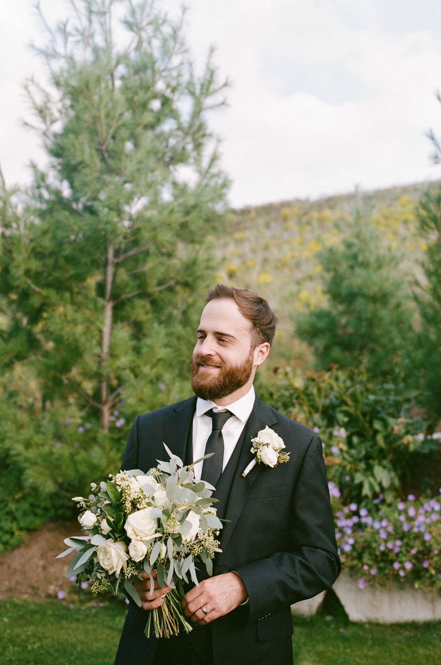 A groom in a black suit holds a delicate white and green wedding bouquet against an outdoor garden backdrop.