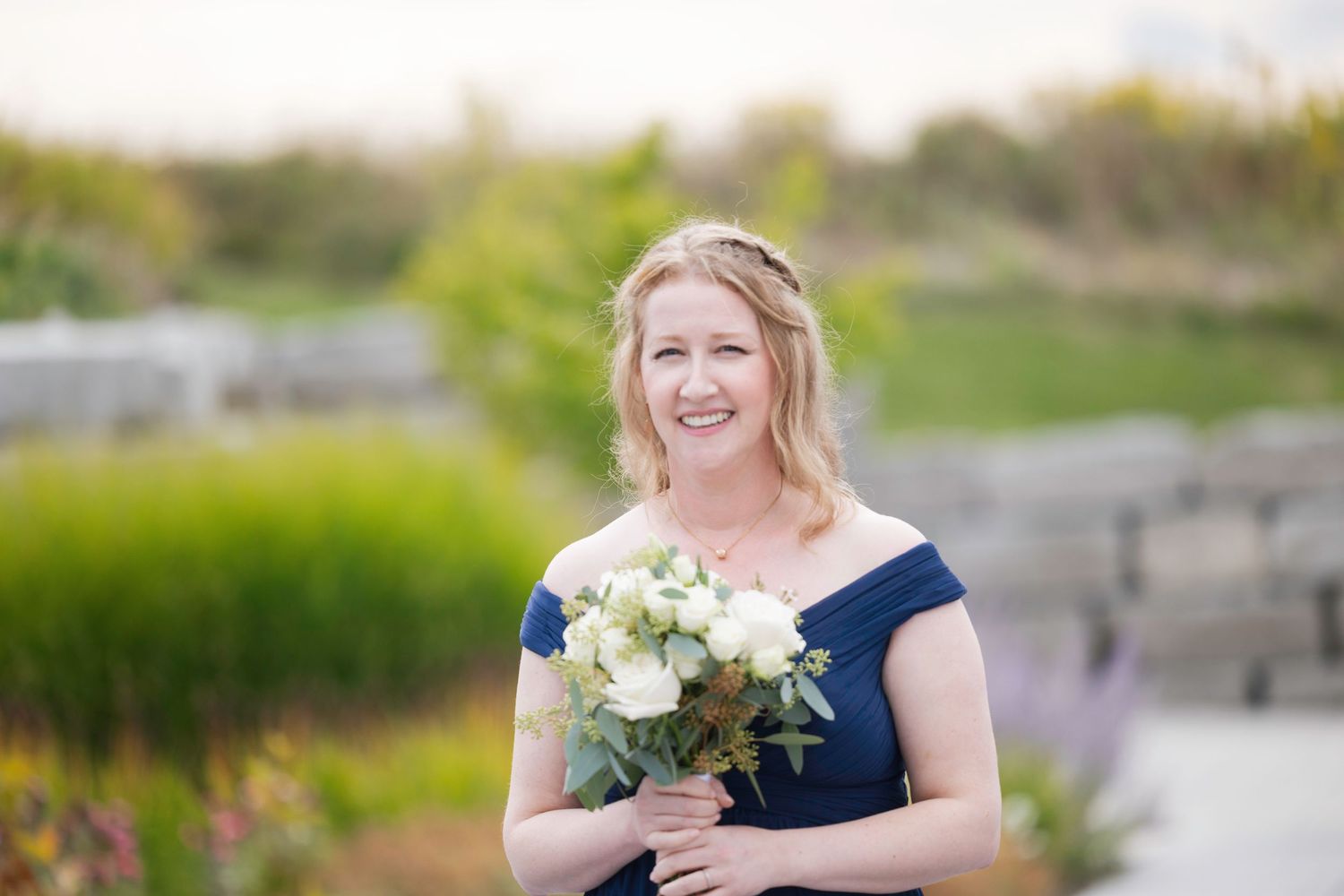 A bridesmaid in a navy blue dress holds a white floral bouquet while standing outdoors on a sunny day.