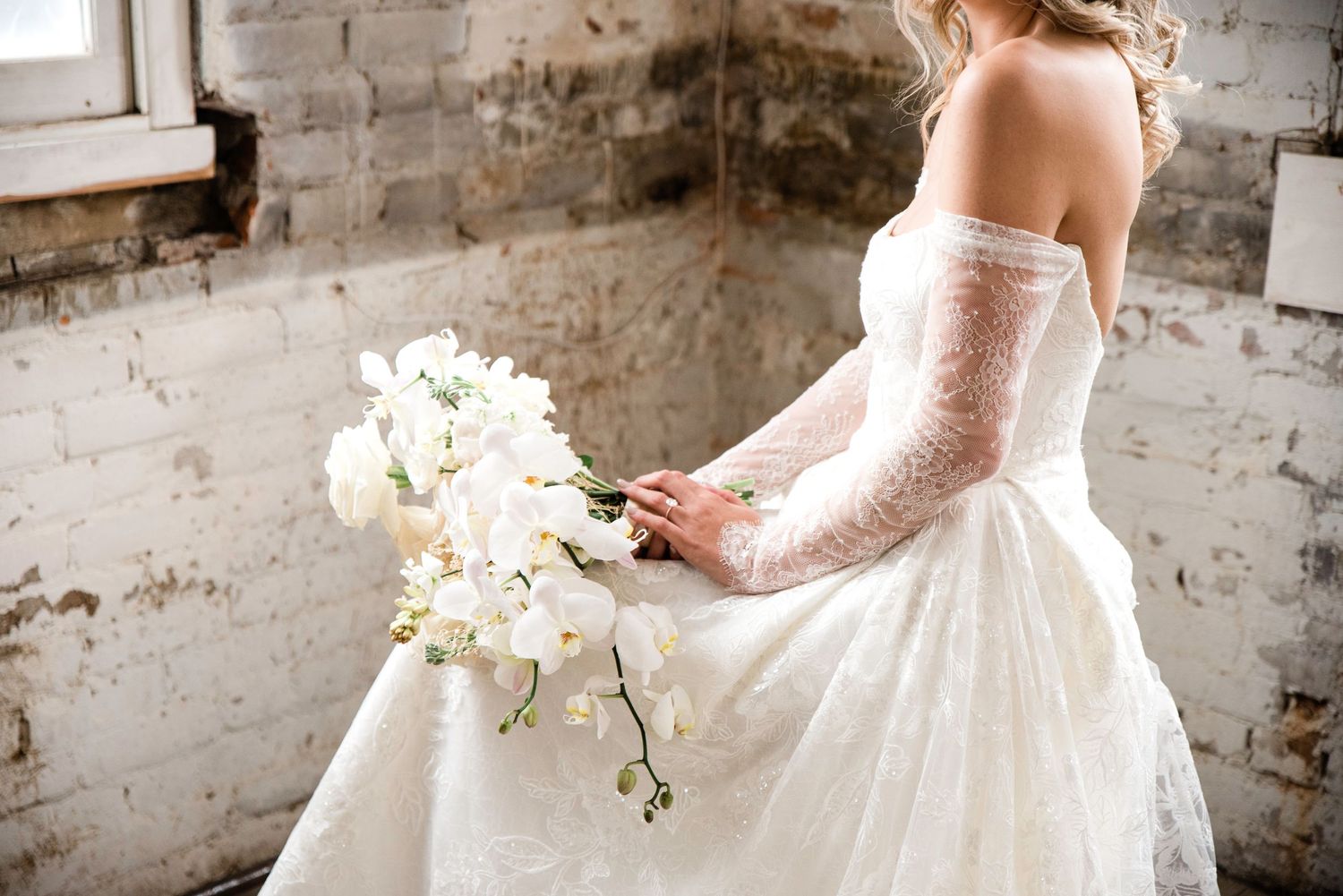 Bride in flowing white gown holds bouquet against exposed brick wall backdrop.