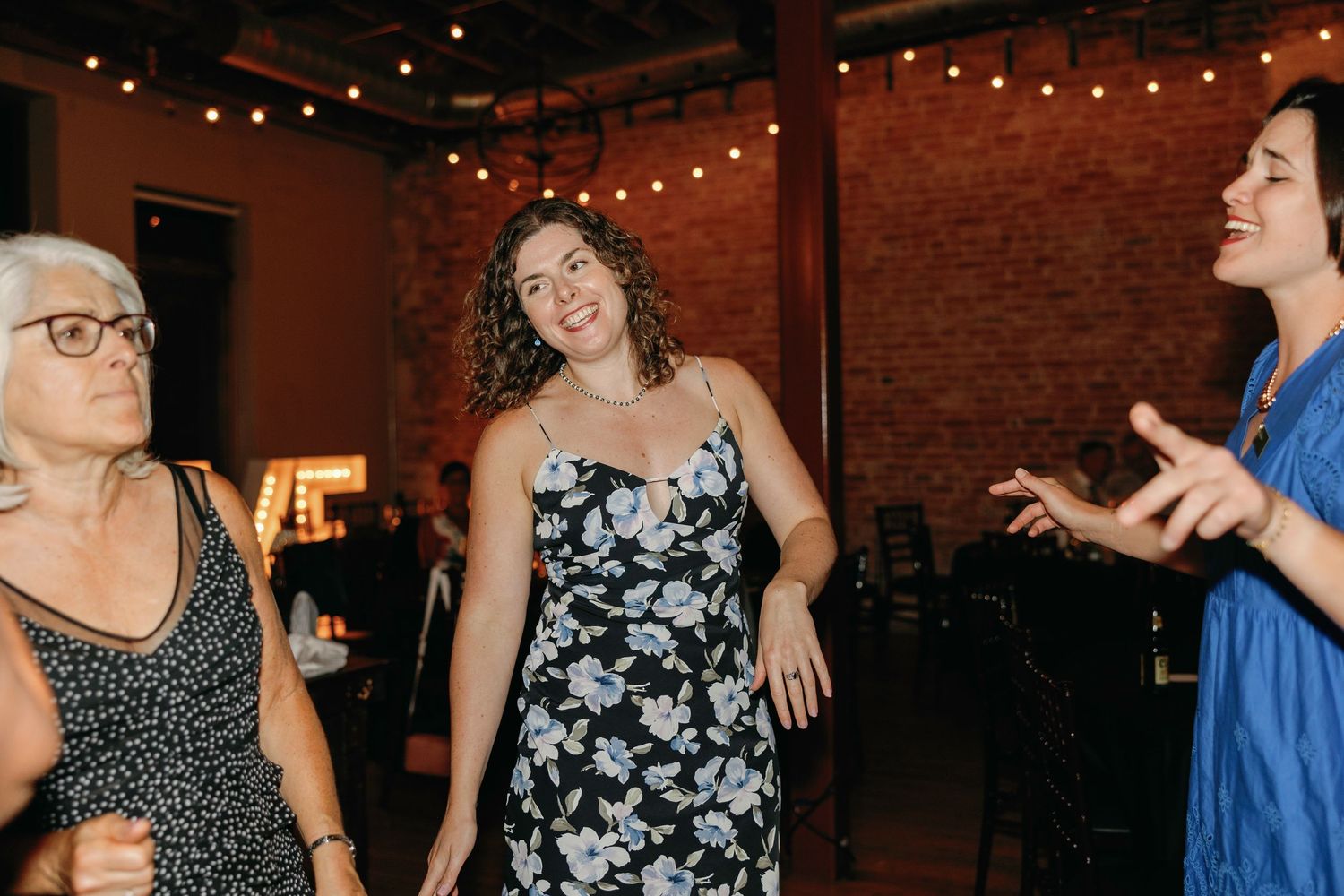 Wedding guests dance and celebrate under warm string lights in a brick-walled reception venue.