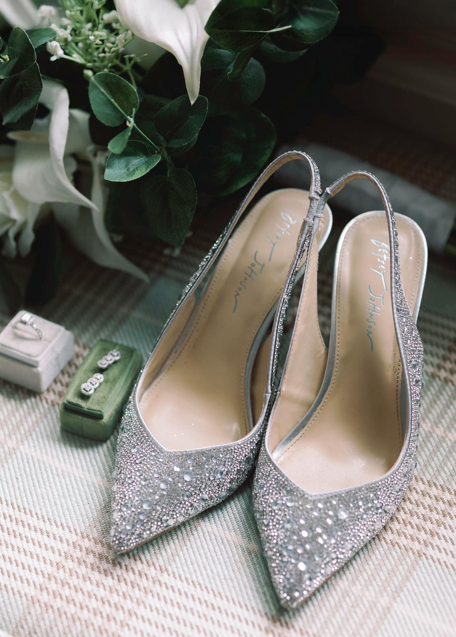 Sparkly silver bridal shoes arranged with greenery on textured carpet.