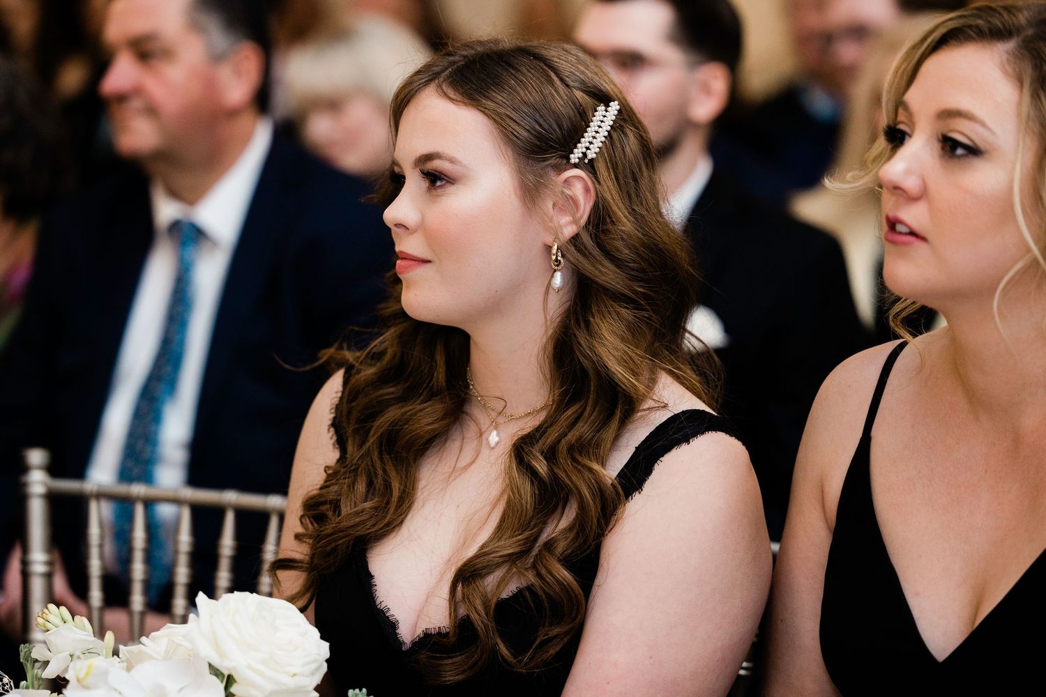 Two women in black dresses seated at a wedding reception, one with wavy hair and a hair clip, holding white flowers.