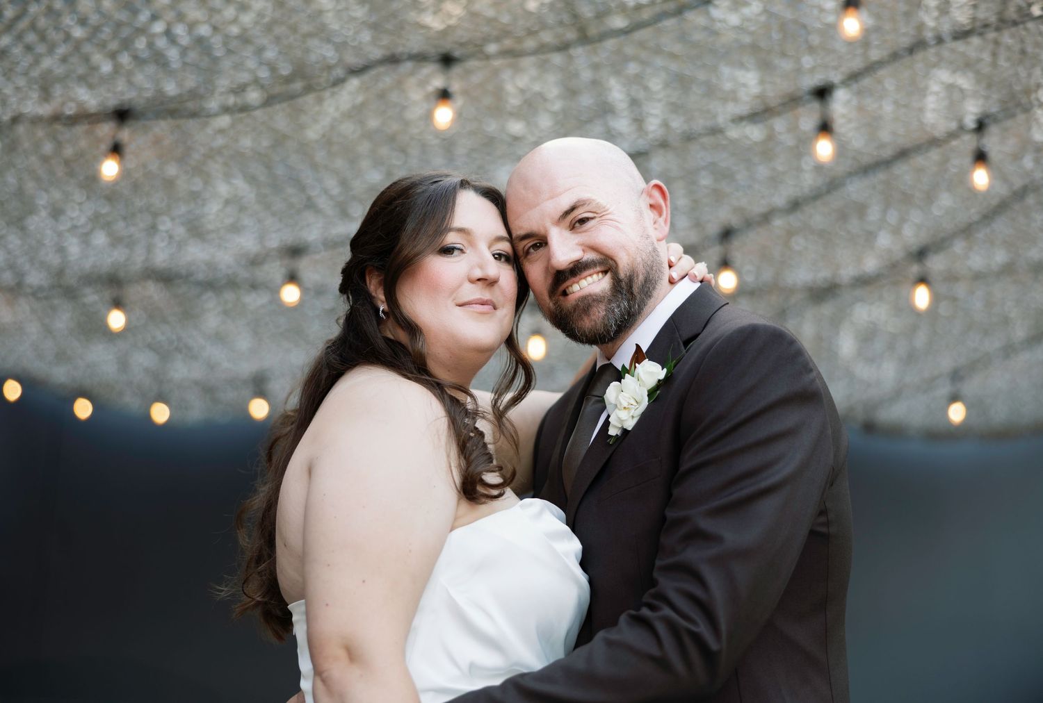Happy newlyweds embrace under string lights, bride in white strapless gown, groom in black suit with boutonniere.