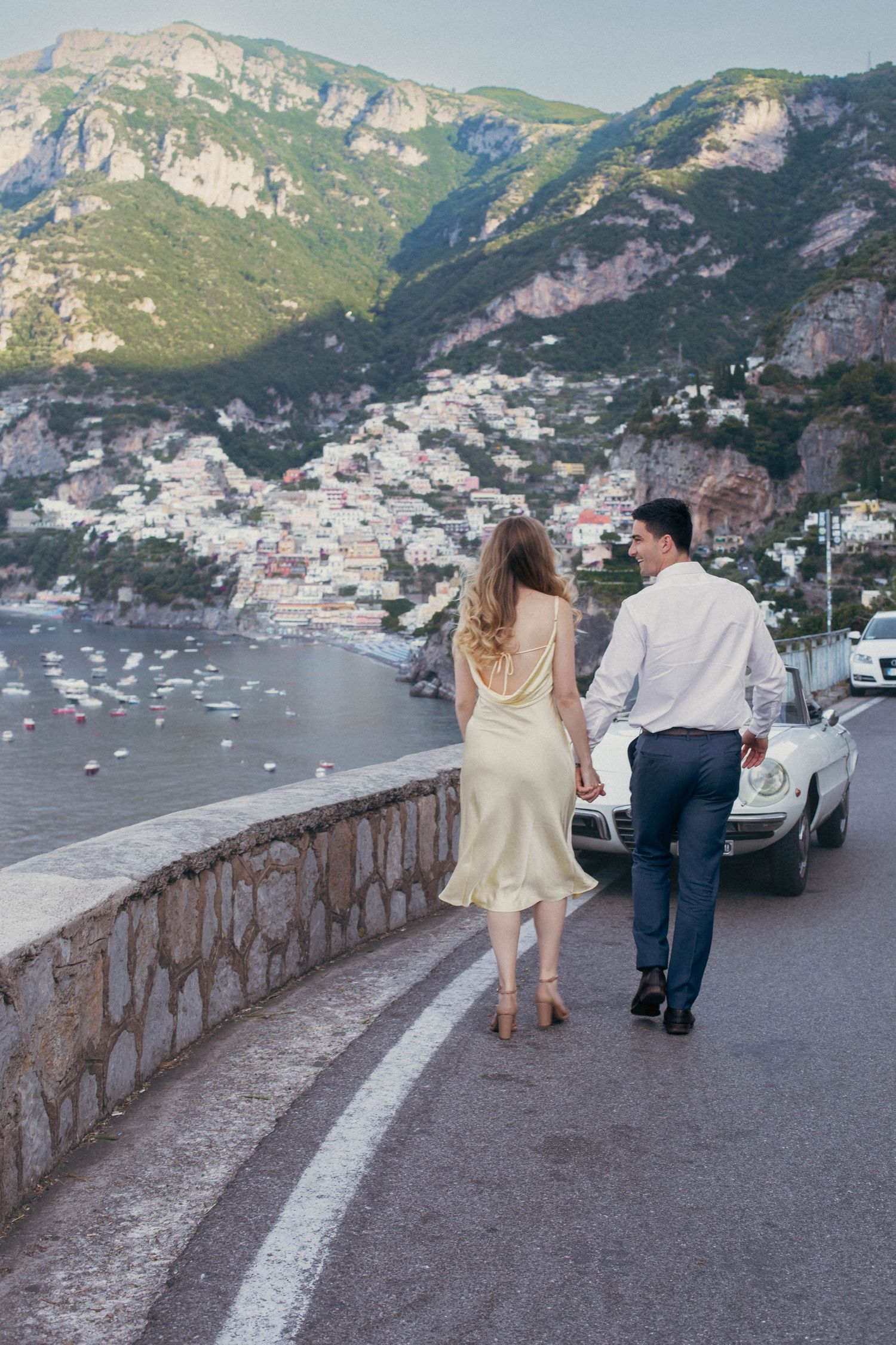 Couple walking hand in hand on Amalfi Coast road with Positano cliffside village and Mediterranean sea in background.